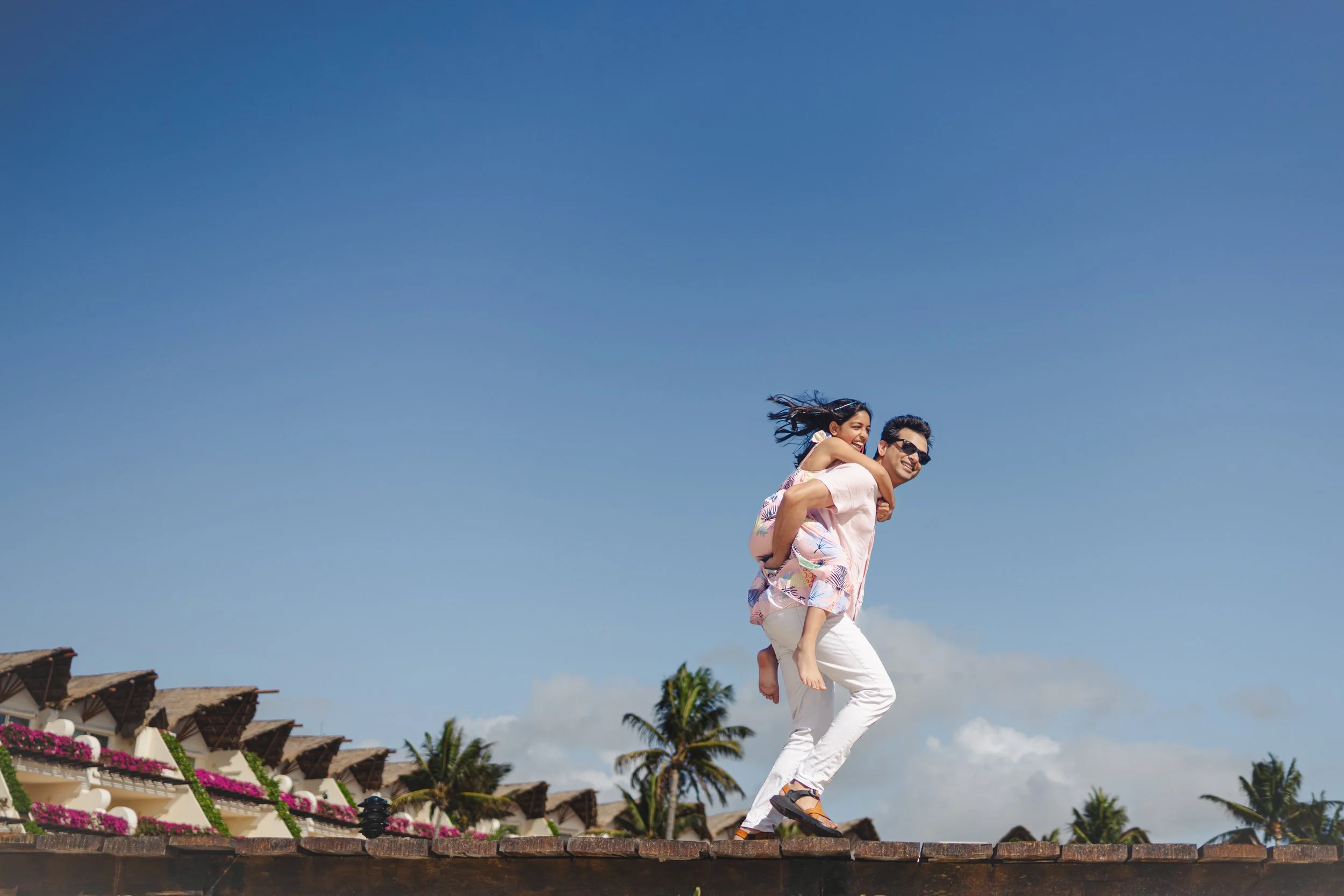 Pareja sonriendo, hombre lleva a una mujer sobre los hombros, al aire libre, con palmeras y casas de techo de paja al fondo, día soleado.