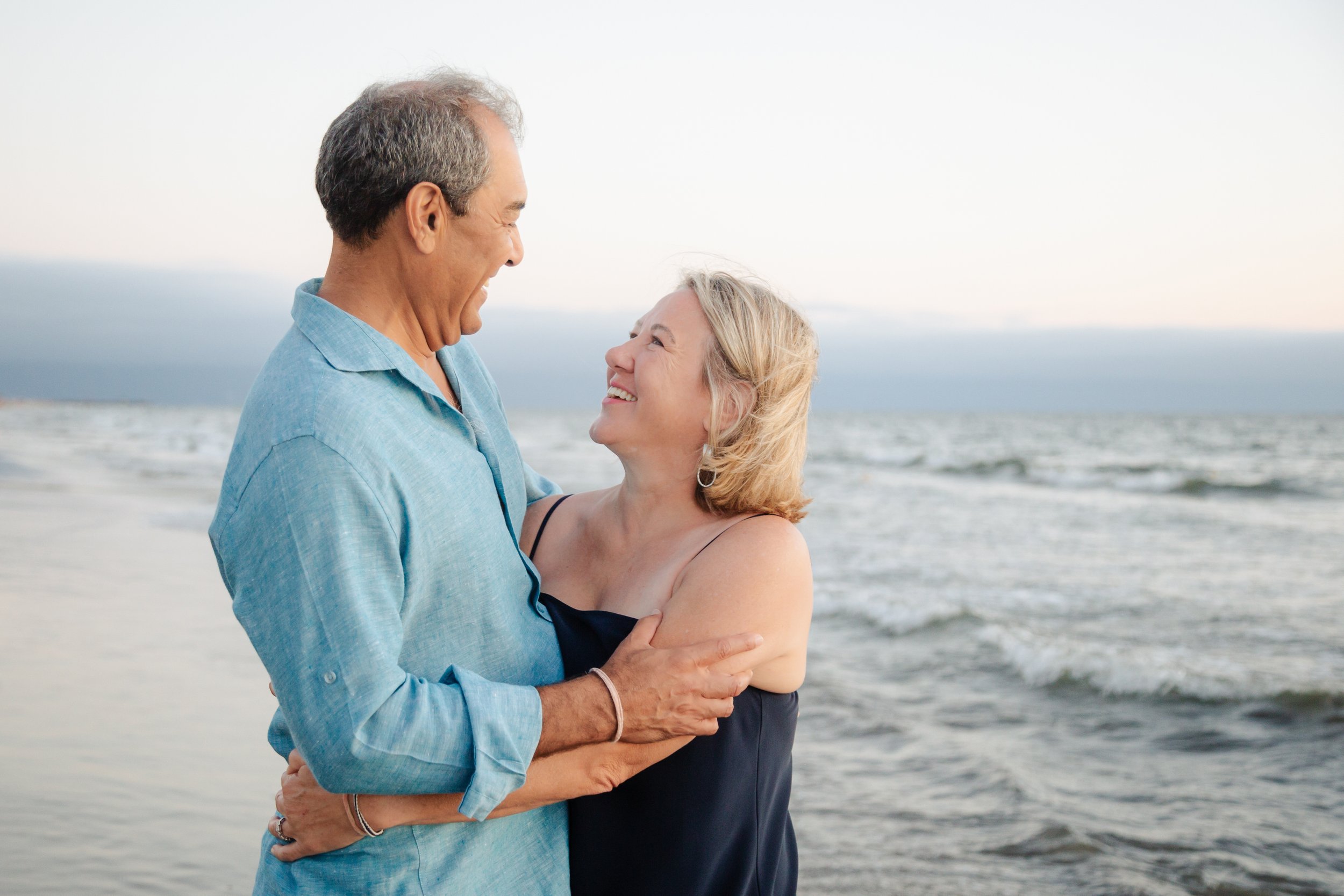 Pareja de ancianos sonriendo y abrazándose en la playa con el mar y el cielo de fondo.