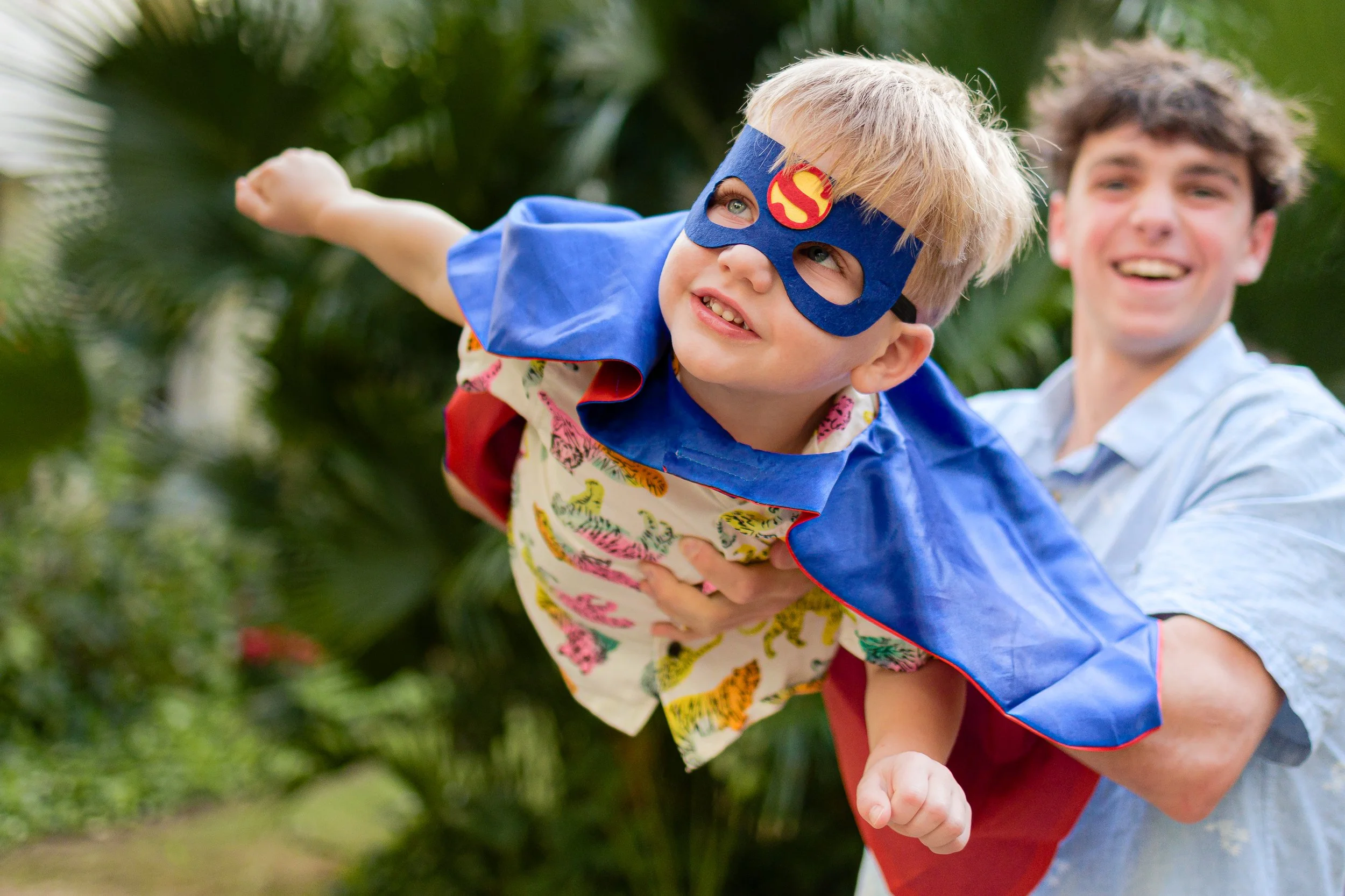 Un niño disfrazado de superhéroe volando mientras un joven le sostiene en una aventura en un entorno natural con vegetación.