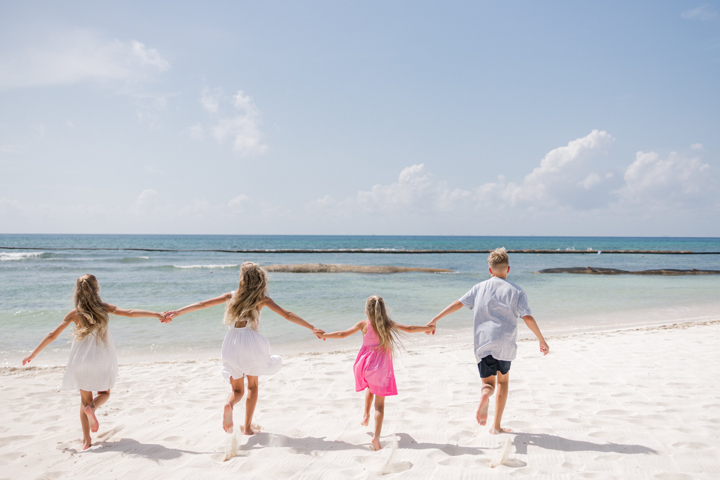 Niños corriendo y bailando en la playa, tomados de las manos, con el mar y el cielo despejado al fondo.