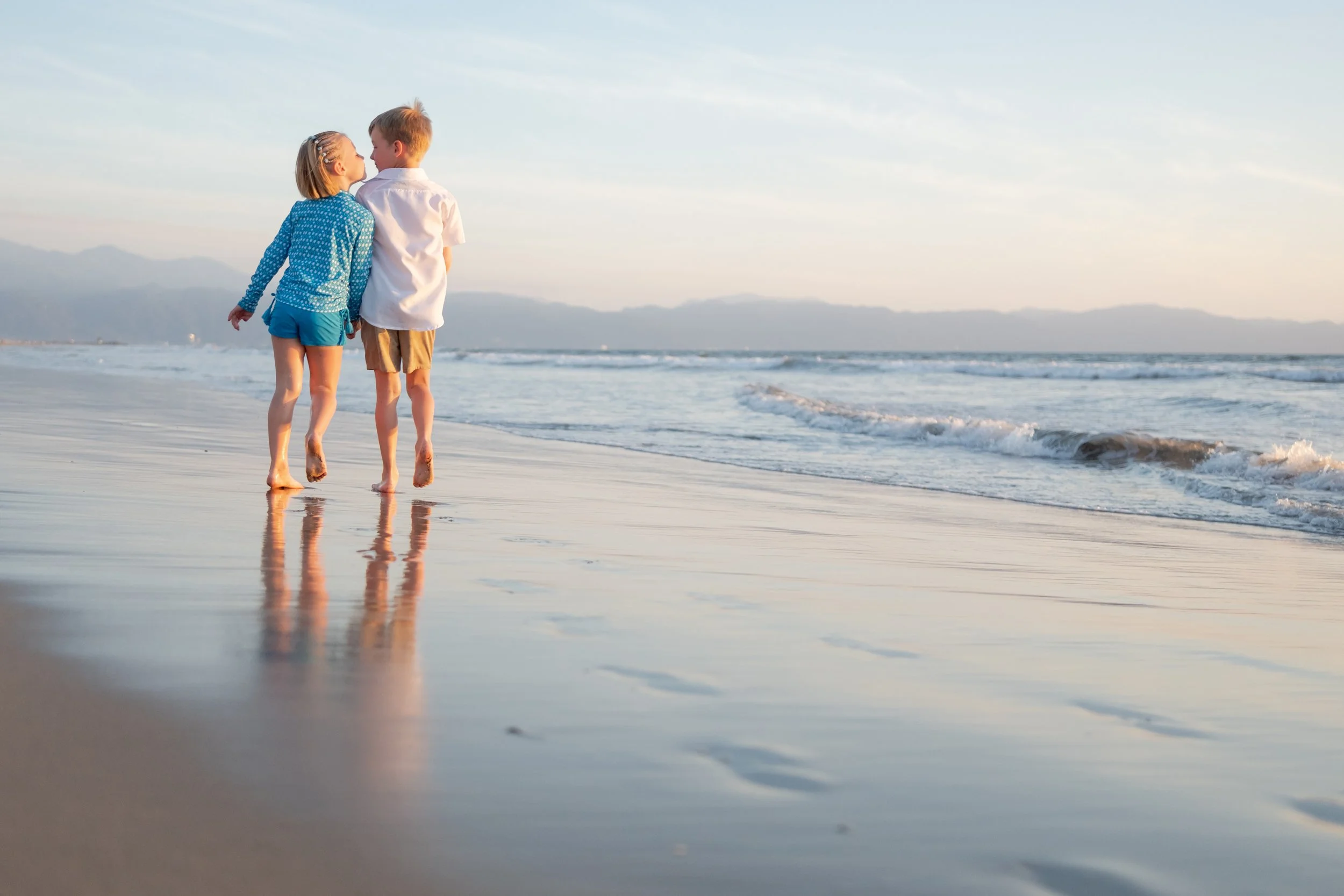 Niños caminando junto a la orilla del mar en la playa, tomados de la mano y aparentando estar en verano.