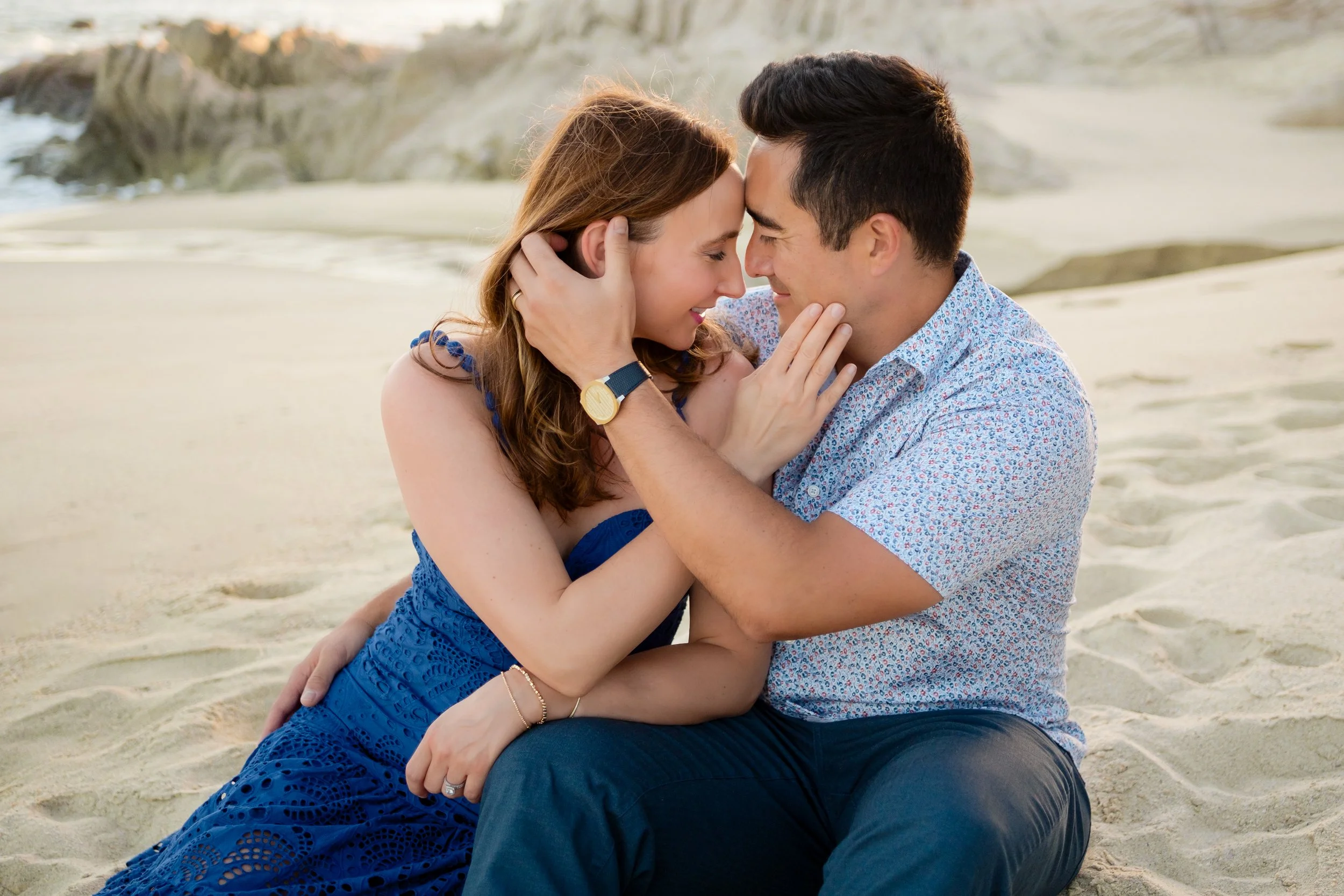 Una pareja joven en la playa, tocándose sus rostros y sonriendo, con el fondo de rocas y arena.