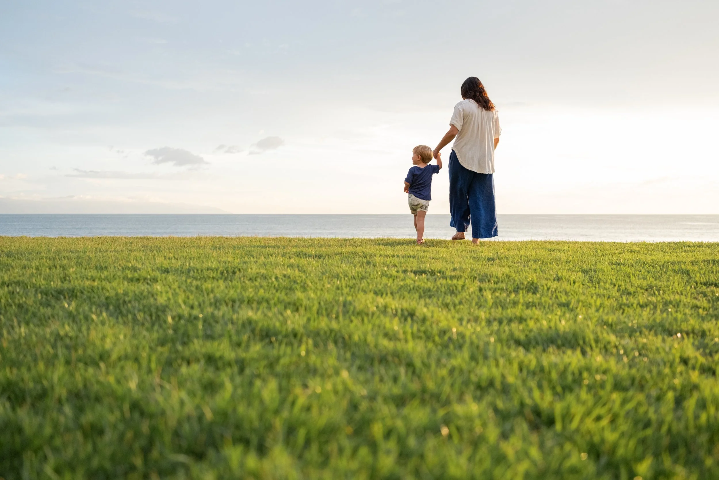 Una mujer con un niño caminan por una pradera frente al mar, en un atardecer soleado.