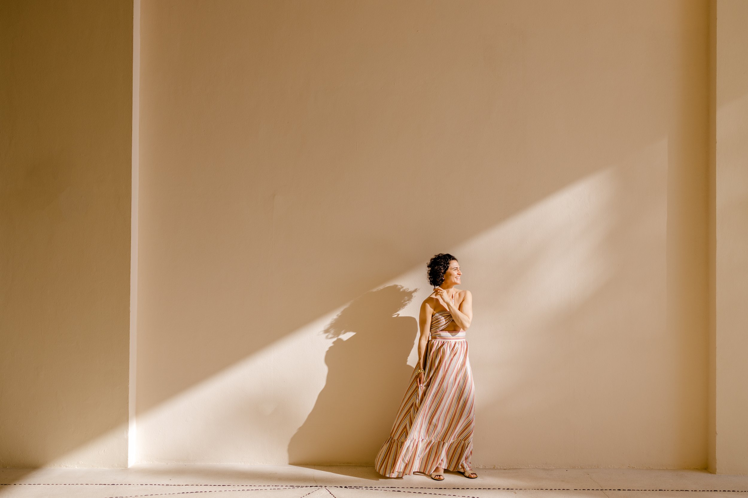Mujer de cabello rizado en un vestido largo con rayas, sonriendo en una pared beige, con sombra y luz brillante.