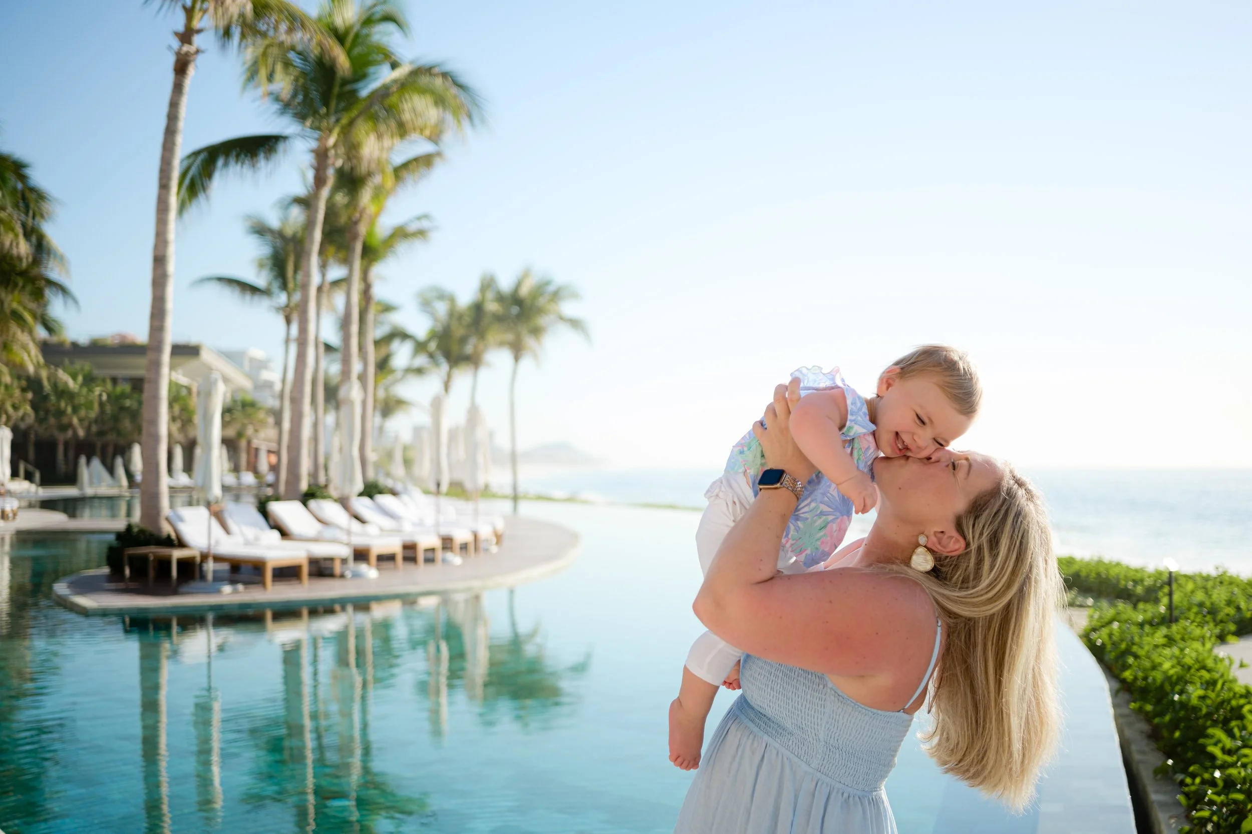 Una mujer con pendientes grandes y cabello rubio sostiene en brazos a un niño pequeño, ambos sonríen y se besan en la mejilla, en un entorno tropical con palmeras y piscina.