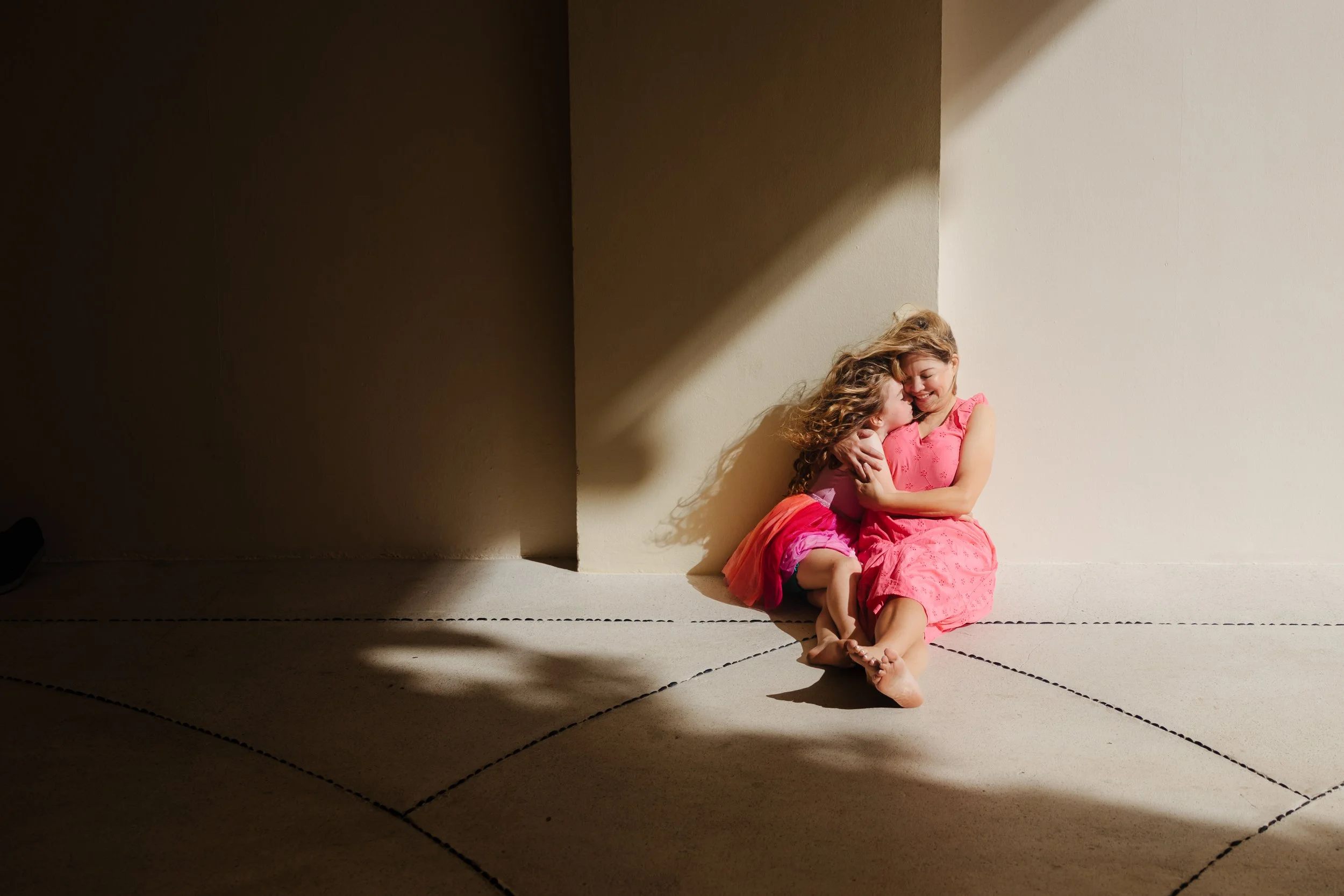 Dos niñas con vestidos rosados, una con el cabello rizado, abrazándose y sonriendo en una esquina iluminada por luz natural.