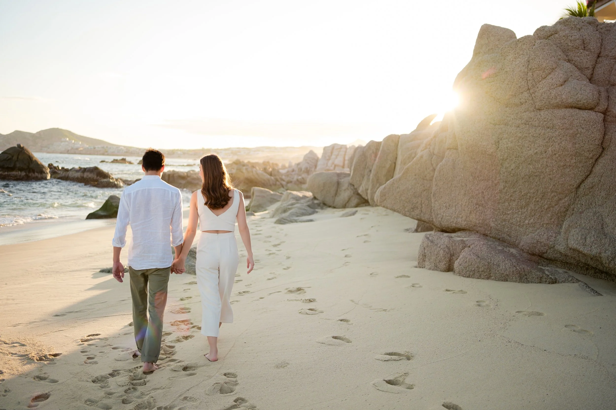una pareja caminando de la mano en una playa durante el atardecer, con rocas grandes a la derecha y arena con huellas