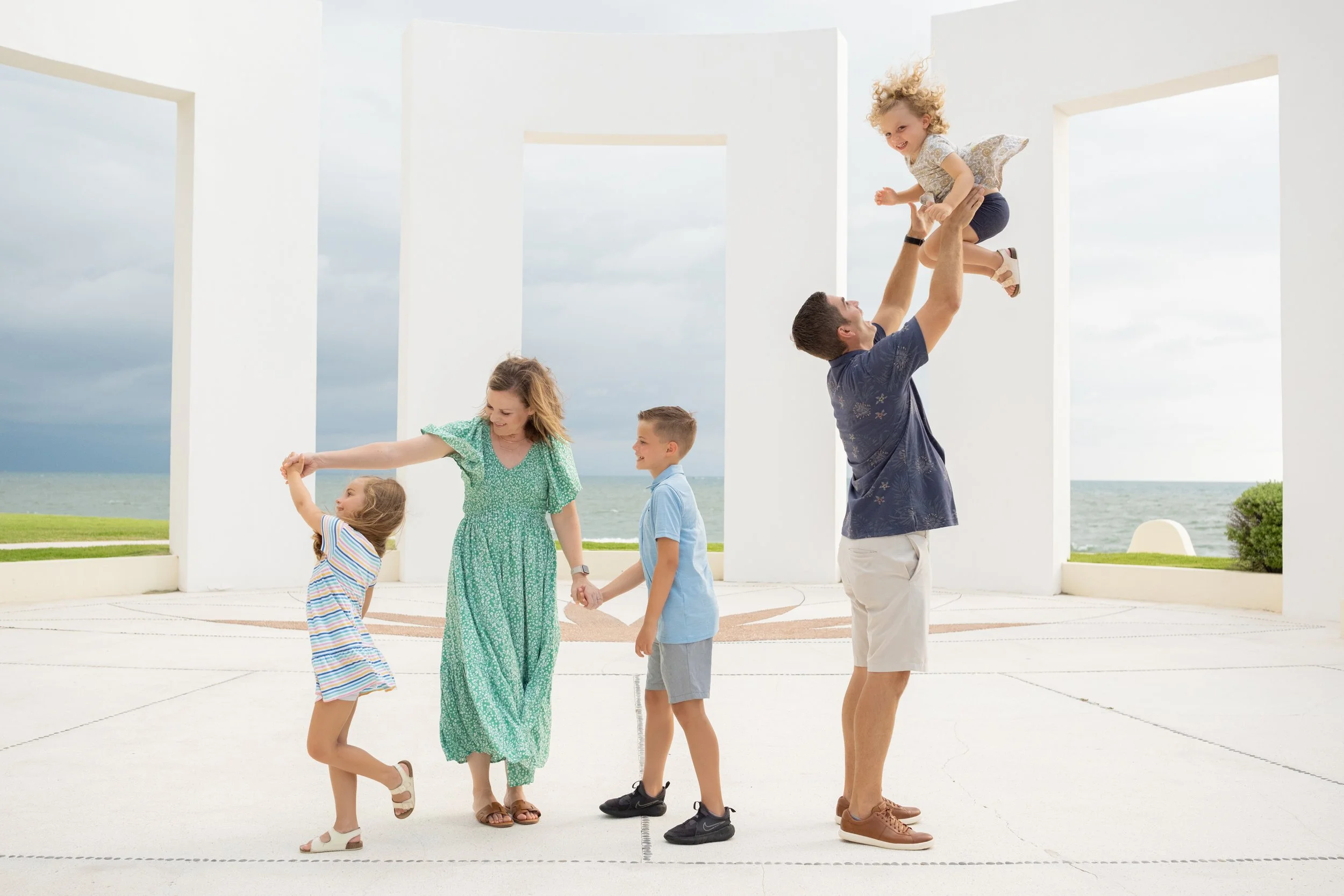familia en la playa jugando, un hombre levanta a una niña mientras los otros niños y una mujer observan y participan en la diversión