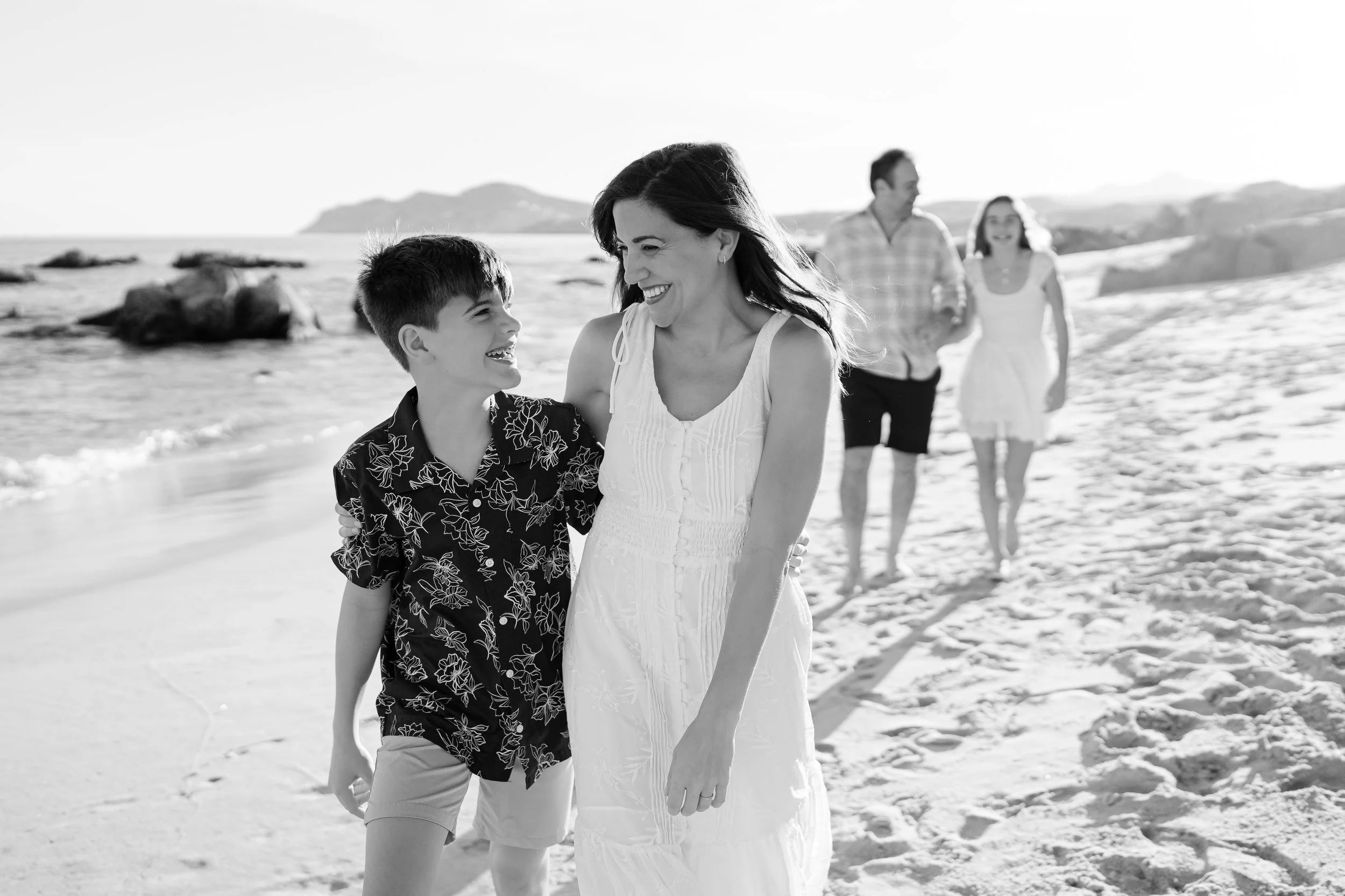 Grupo familiar en la playa, caminando y sonriendo, en un día soleado, con rocas y mar al fondo.