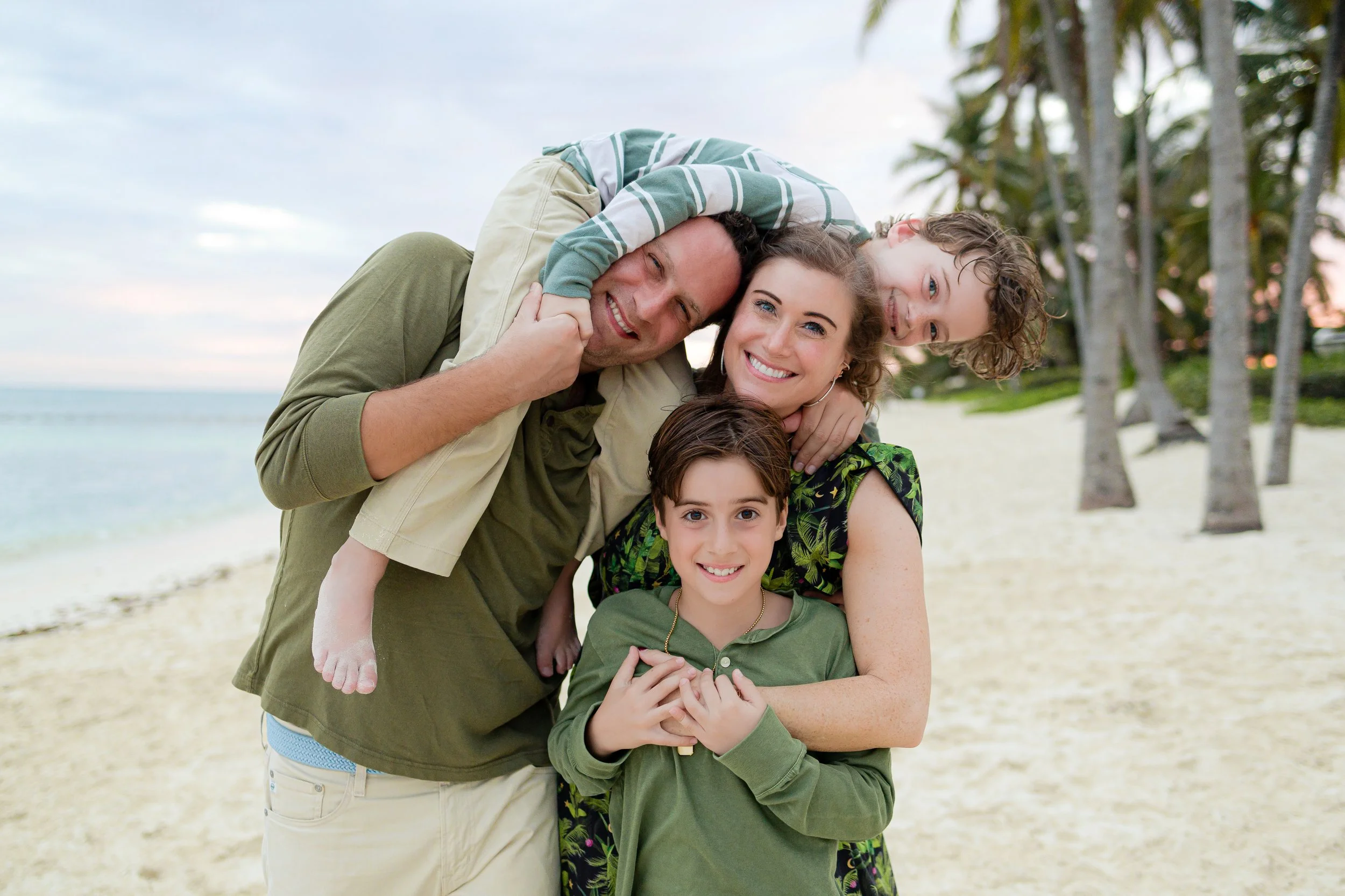 Familia disfrutando en la playa con palmeras al atardecer, sonriendo y abrazándose.