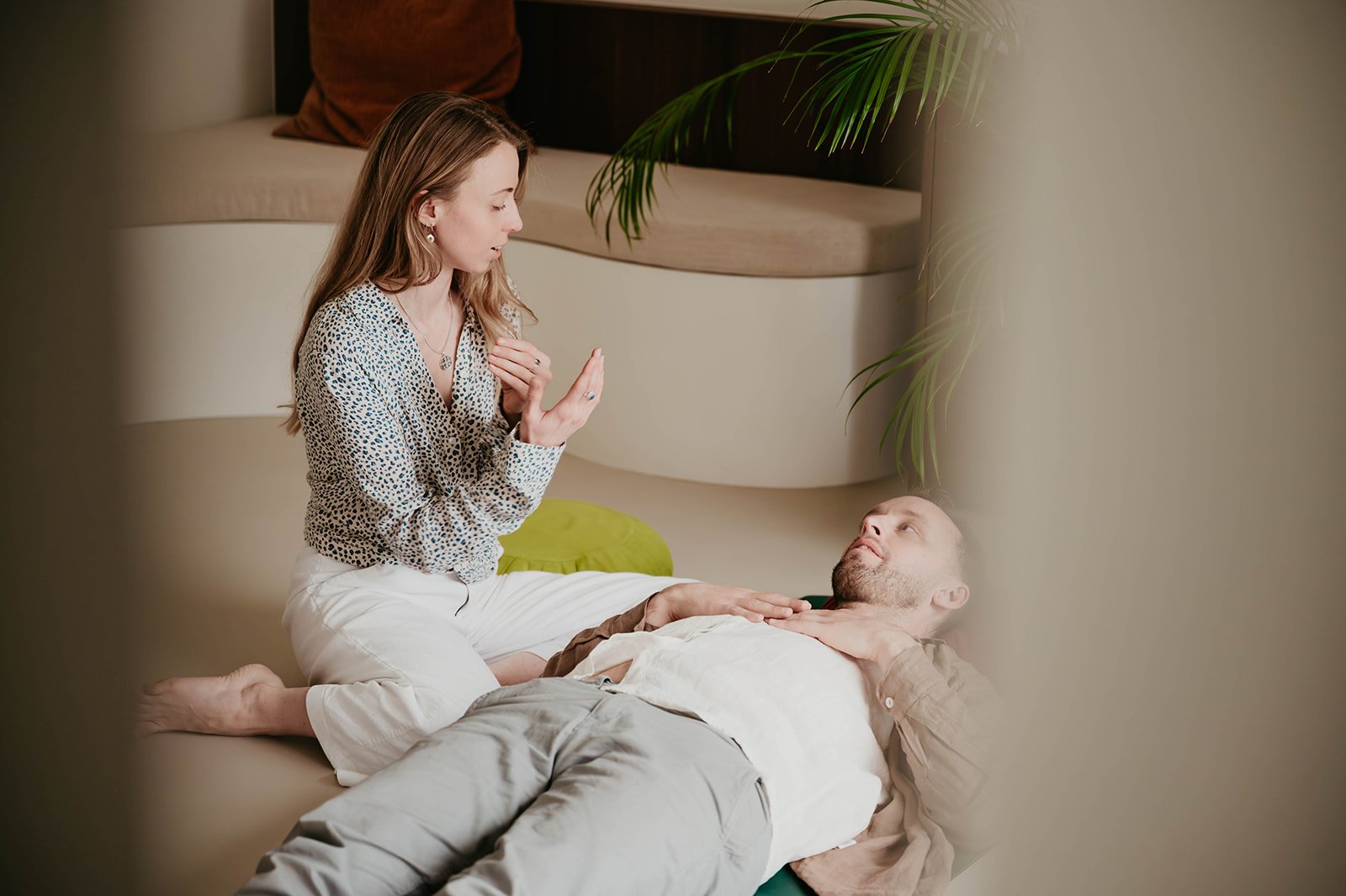 A woman kneeling on a couch talking to a man lying on the floor, touching his chest. There is a plant and cushions in the background.