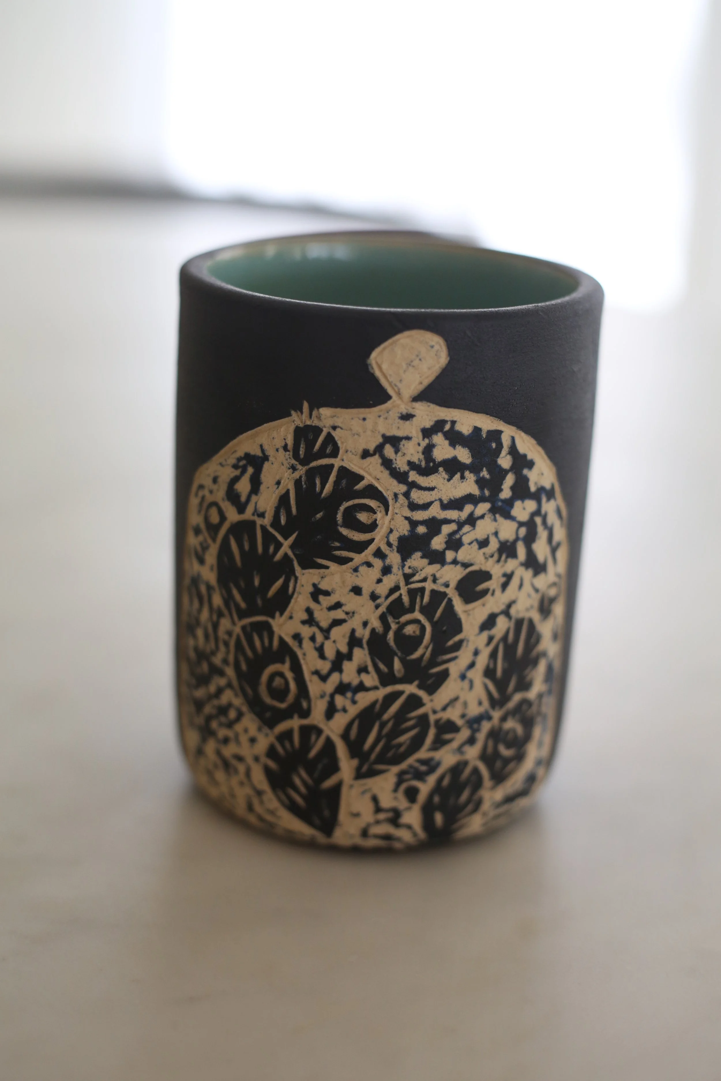 A ceramic tumbler by Amy Ambrose with sgraffito art of a prickly pear in a dome.