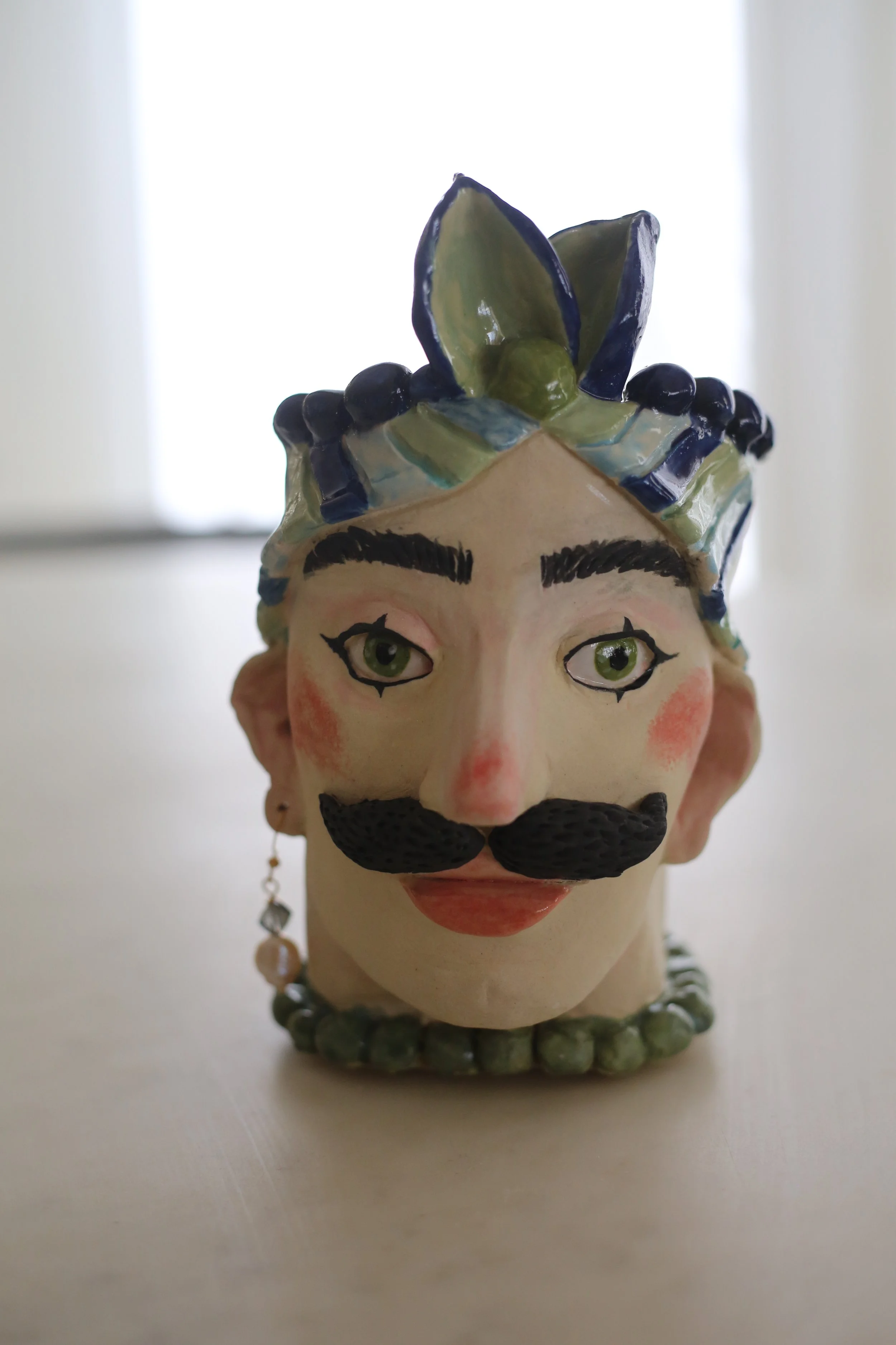 "Guillermo" Ceramic Sicilian Head by Amy Ambrose. A man with a moustache, wearing a colourful headdress, with large green eyes and an earring, set against a plain background.
