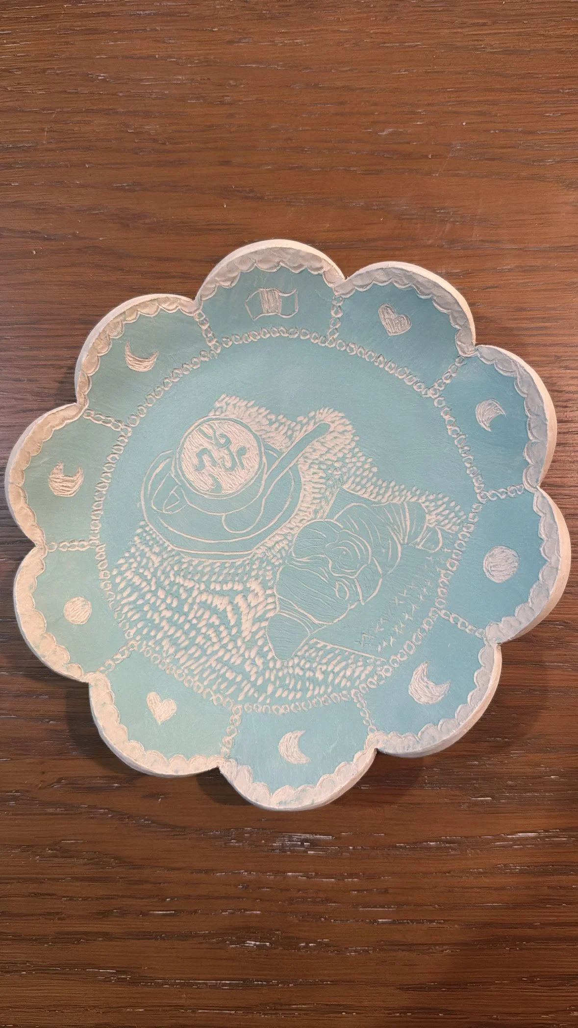 A decorative ceramic plate with a light blue background, featuring an illustration of a croissant and a coffee with music on it, surrounded by celestial motifs such as moons, stars, and hearts.