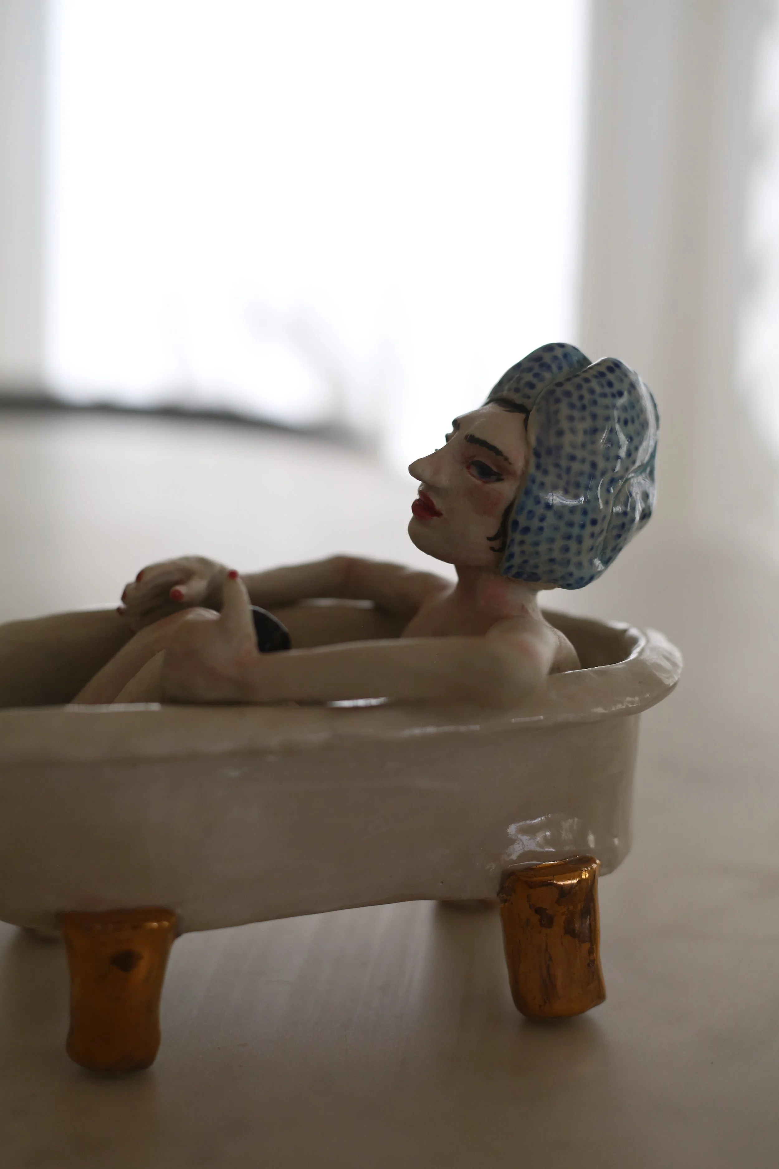 A ceramic figurine by Amy Ambrose of a woman with a headscarf, sitting inside a bath with gold lustre accents, holding a small black matcha cup, with tiny beads inside the bath. Other side view.