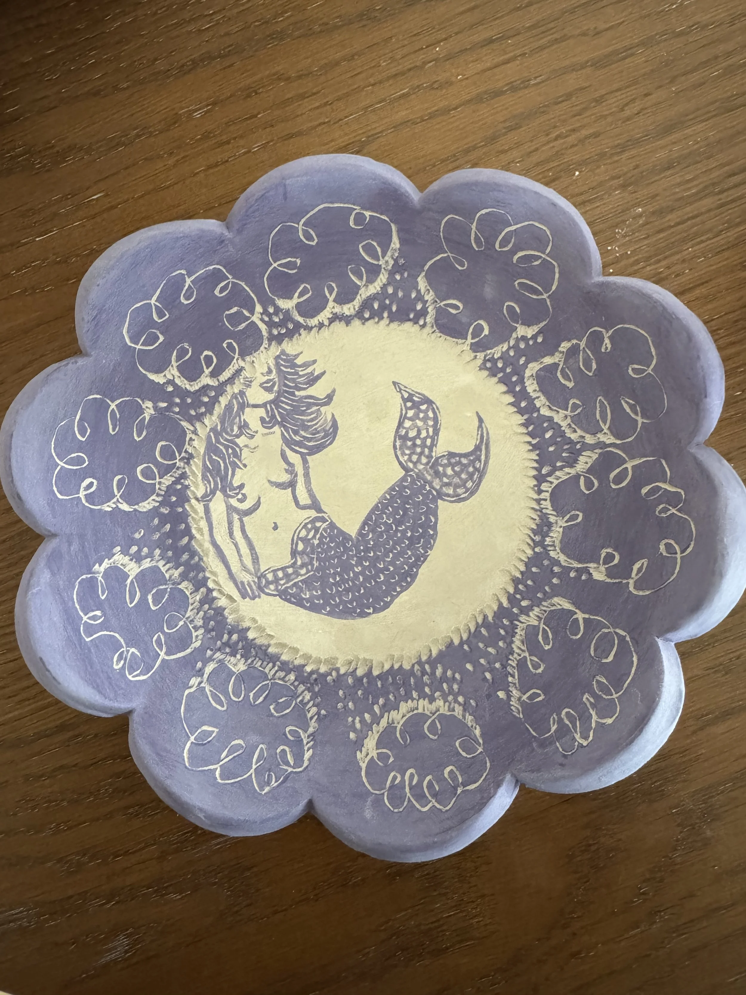 "Unbothered by the rain because I live in the sea". A scalloped ceramic plate featuring sgraffito illustrations of rain clouds and a mermaid.