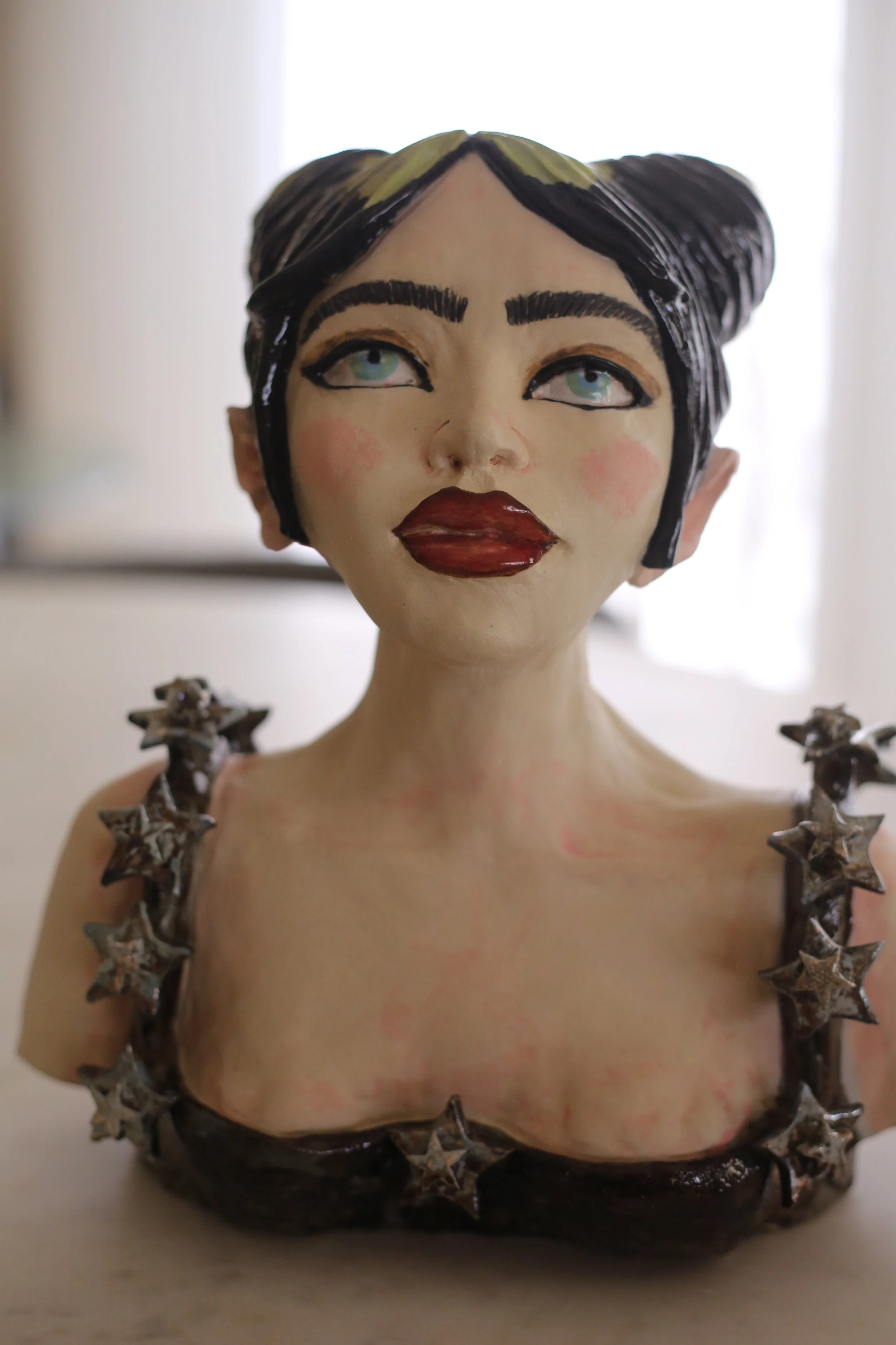 "What was I made for?" Ceramic bust by Amy Ambrose inspired by Billie Eilish with painted features, wearing a dark dress with star-shaped metallic decorations on the straps.