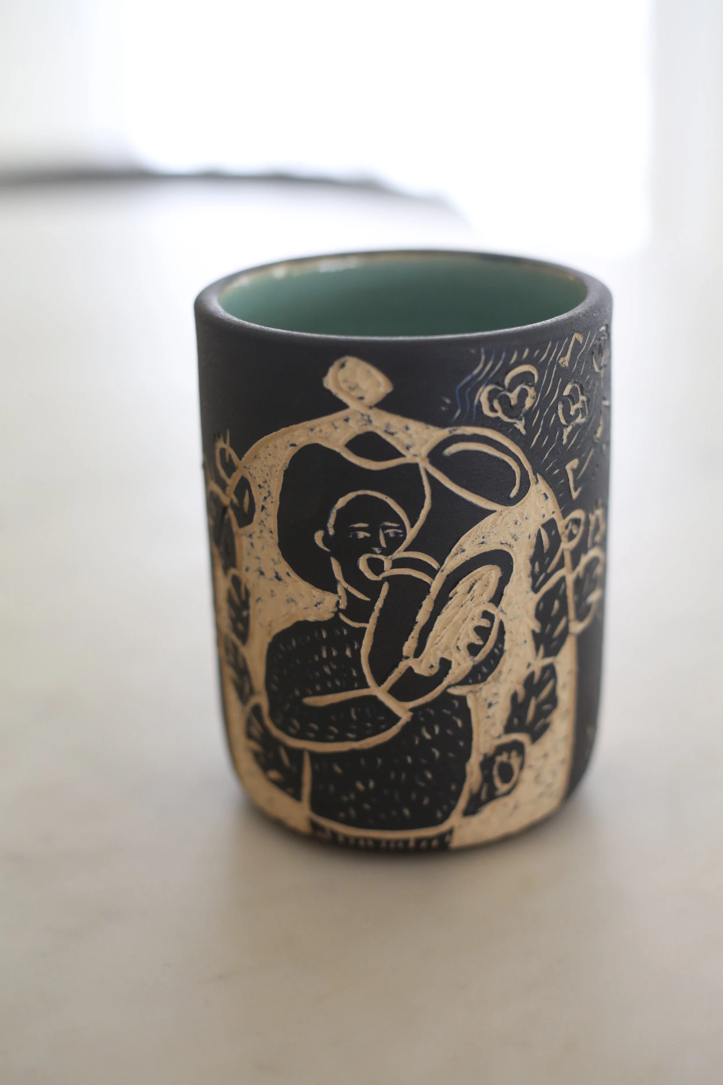 A ceramic tumbler by Amy Ambrose with sgraffito art of a man playing a trumpet in a dome. 