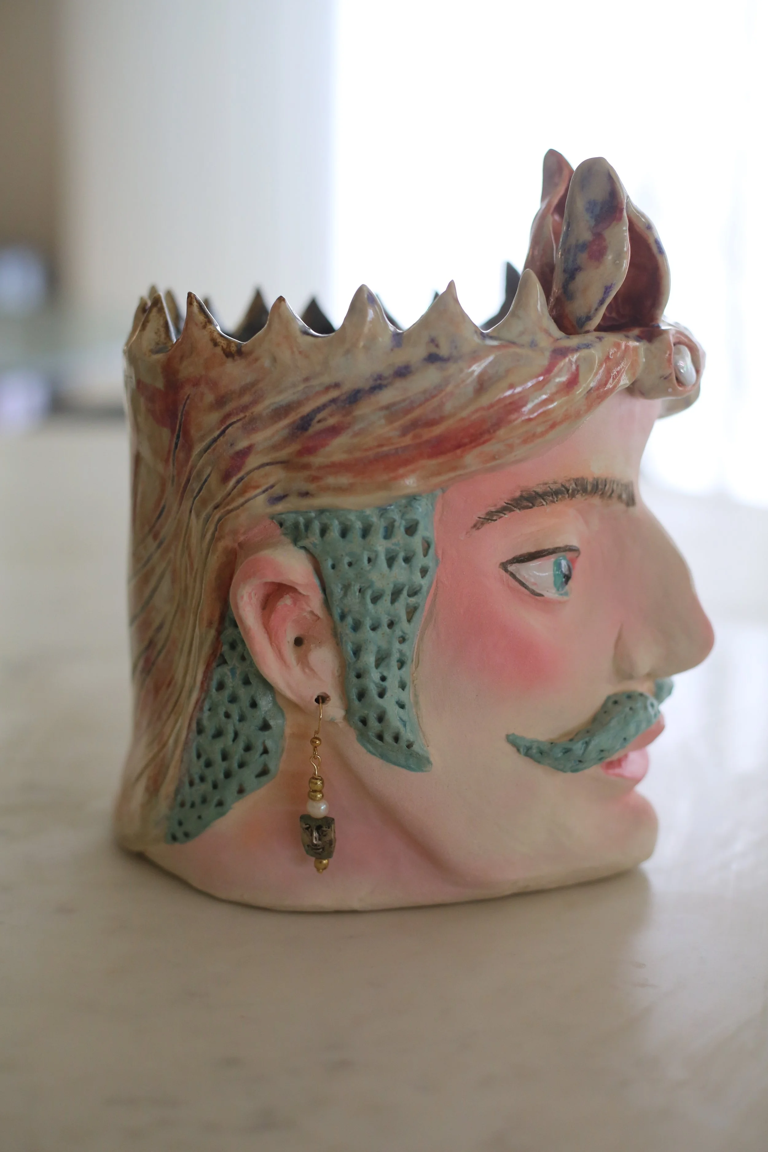 "Guillermo" Ceramic Sicilian Head by Amy Ambrose. Side view. A detailed ceramic sculpture of a human face with an elaborate, textured headdress resembling mountain peaks.