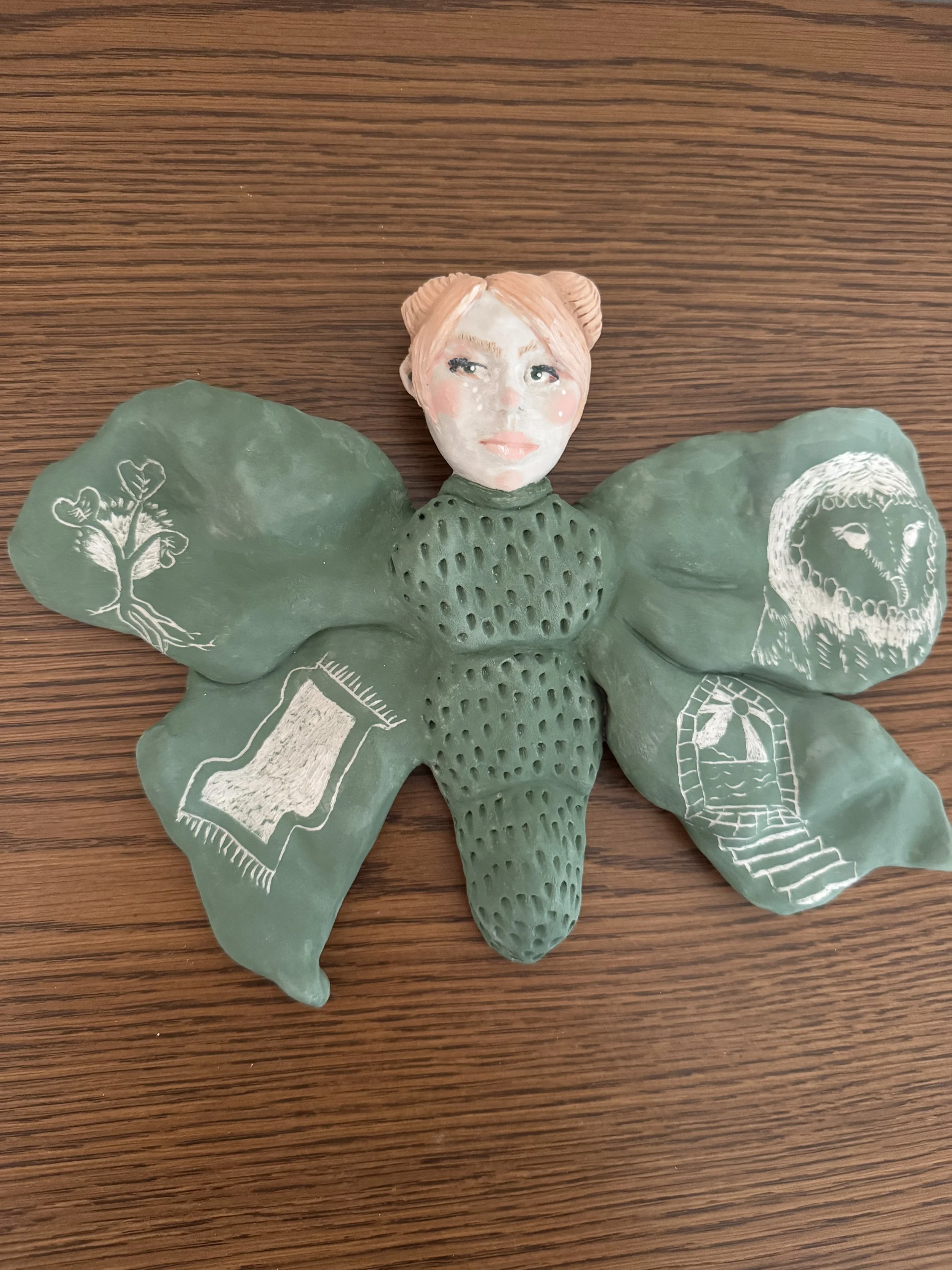 "Growing pains". A ceramic sculpture of a moth woman. Sgraffito illustrations on the wings include an owl, doorway with stairs, growing seedling and a magic carpet.