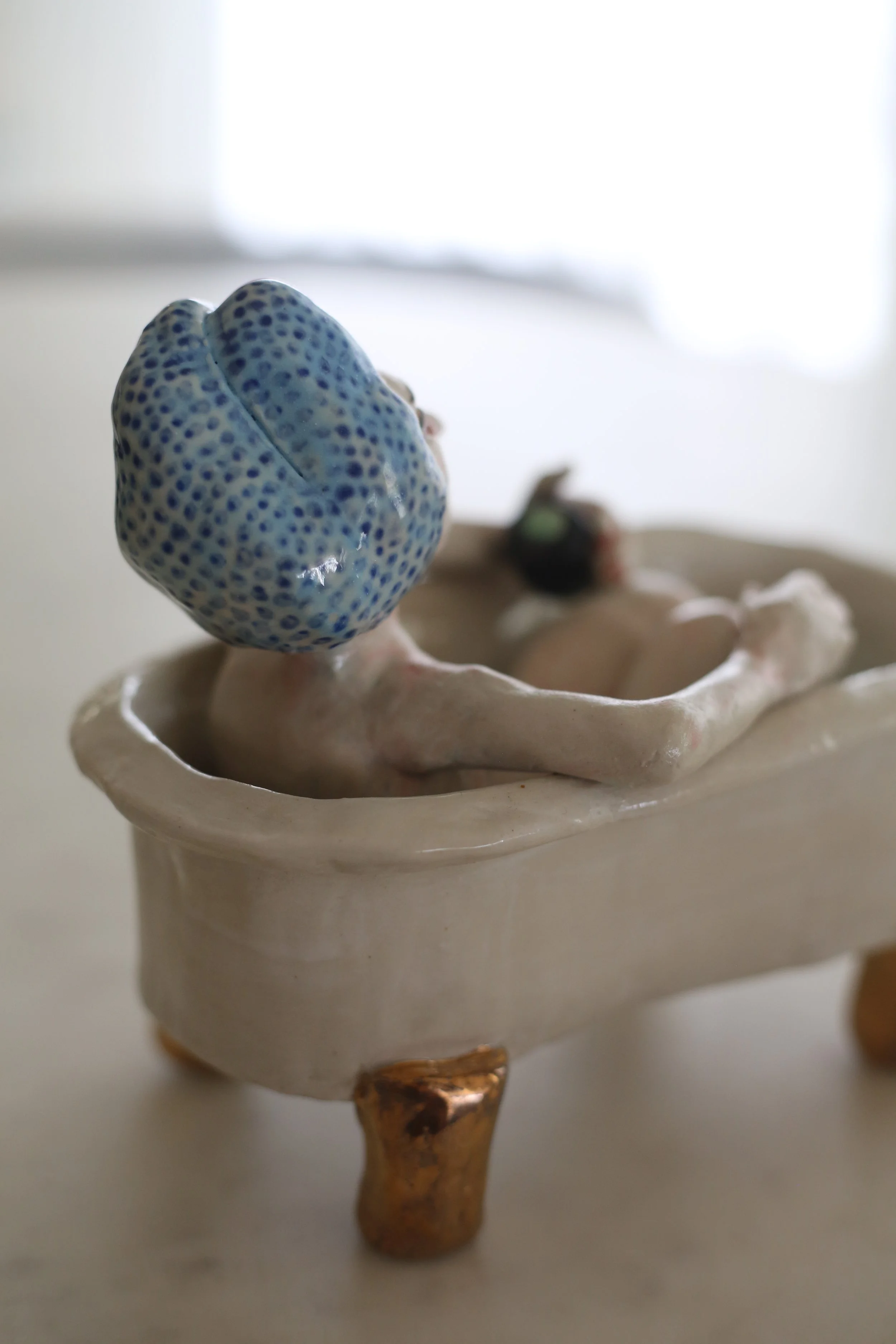A ceramic figurine by Amy Ambrose of a woman with a headscarf, sitting inside a bath with gold lustre accents, holding a small black matcha cup, with tiny beads inside the bath. Back view.