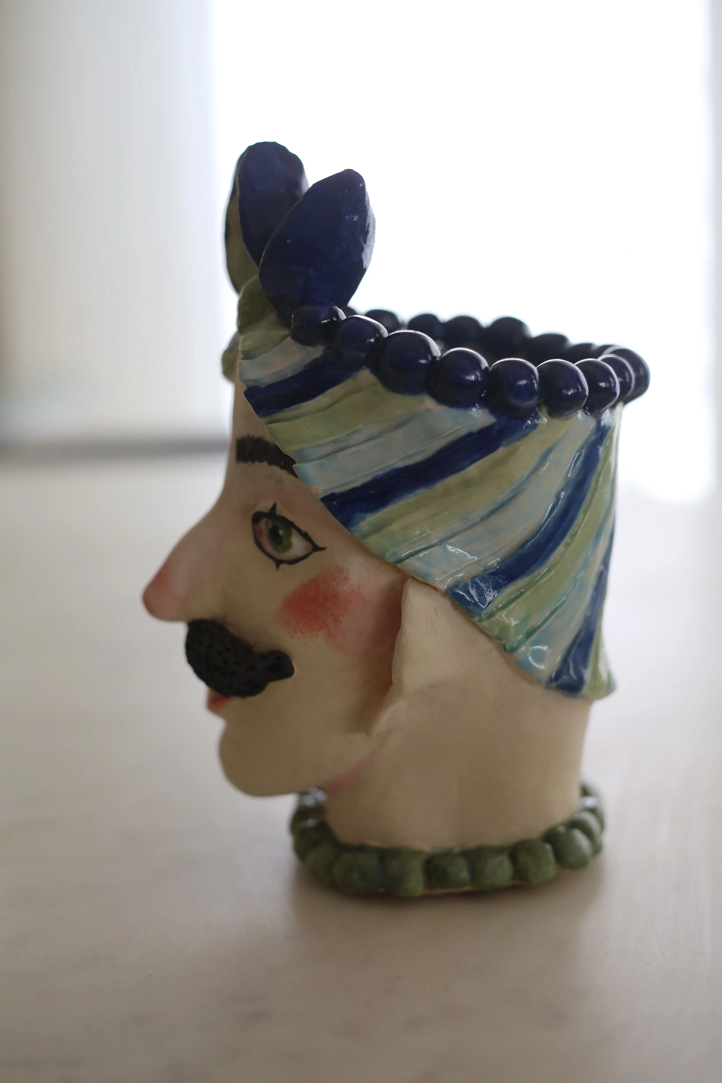 "Guillermo" Ceramic Sicilian Head by Amy Ambrose. A man with a moustache, wearing a colourful headdress, with large green eyes and an earring, set against a plain background. Side View.
