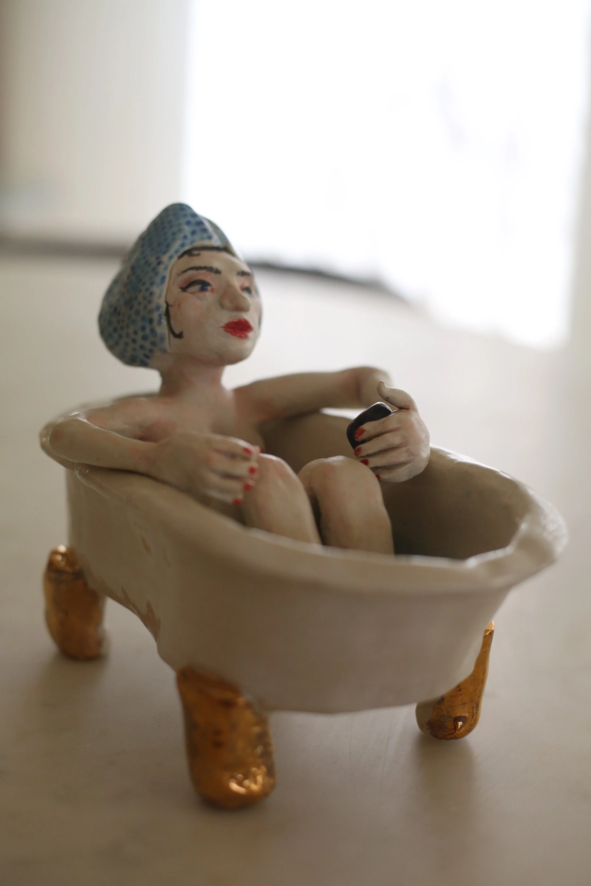 A ceramic figurine by Amy Ambrose of a woman with a headscarf, sitting inside a bath with gold lustre accents, holding a small black matcha cup, with tiny beads inside the bath. Side view.