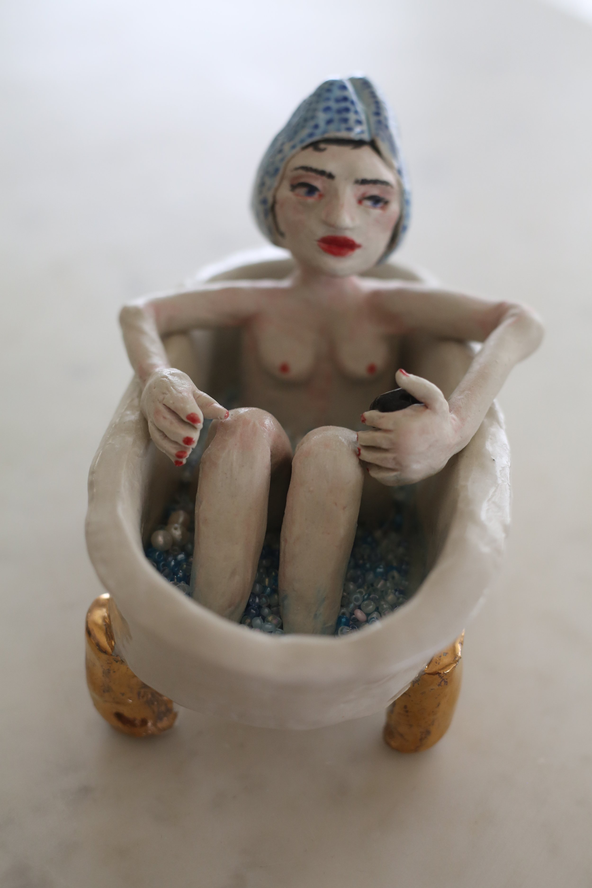 A ceramic figurine by Amy Ambrose of a woman with a headscarf, sitting inside a bath with gold lustre accents, holding a small black matcha cup, with tiny beads inside the bath.
