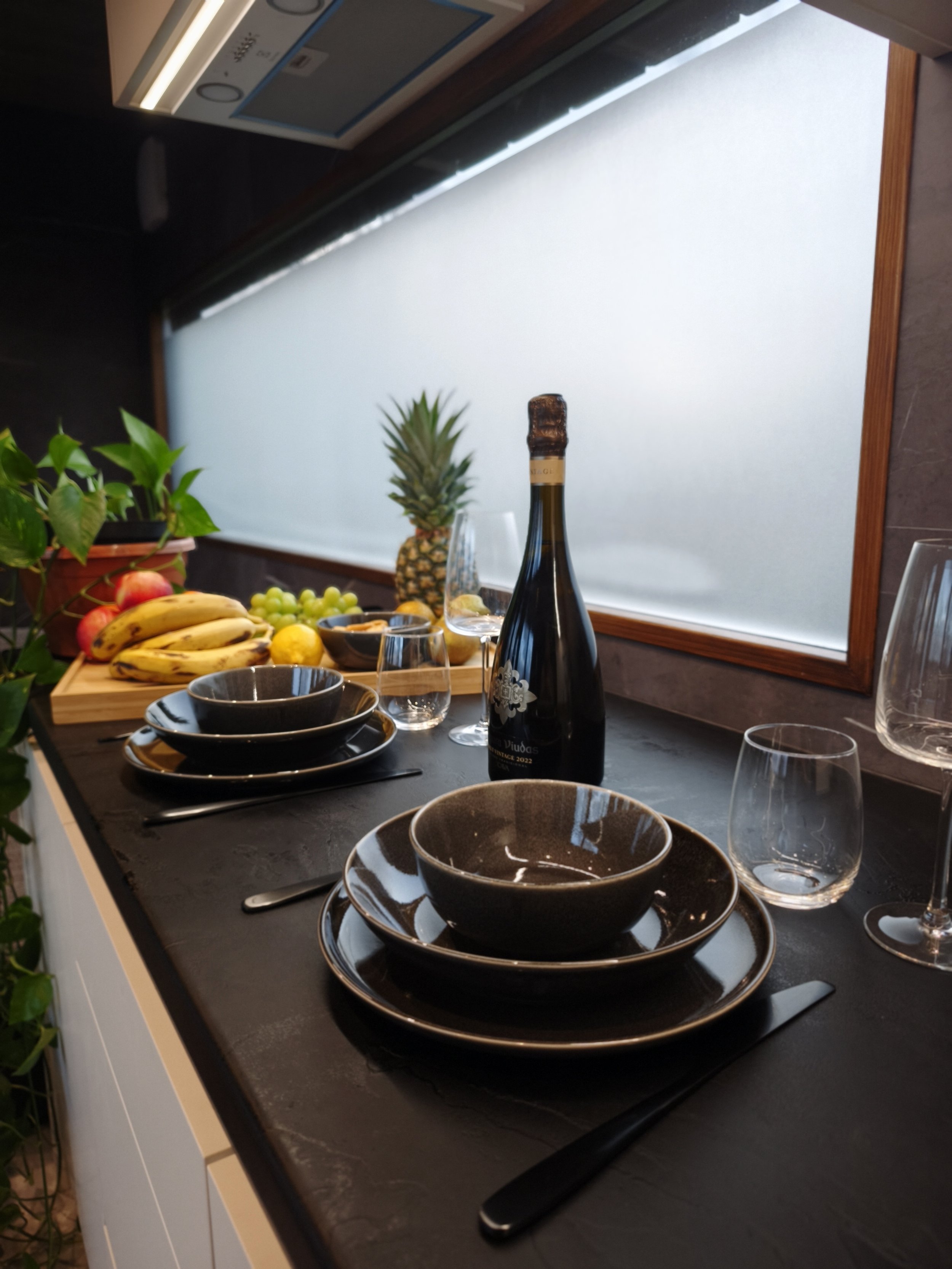 A table set with black dishes, glasses, and utensils, with a bottle of wine, fresh fruit including bananas, apples, grapes, and a pineapple, and a window with a white blind in the background.