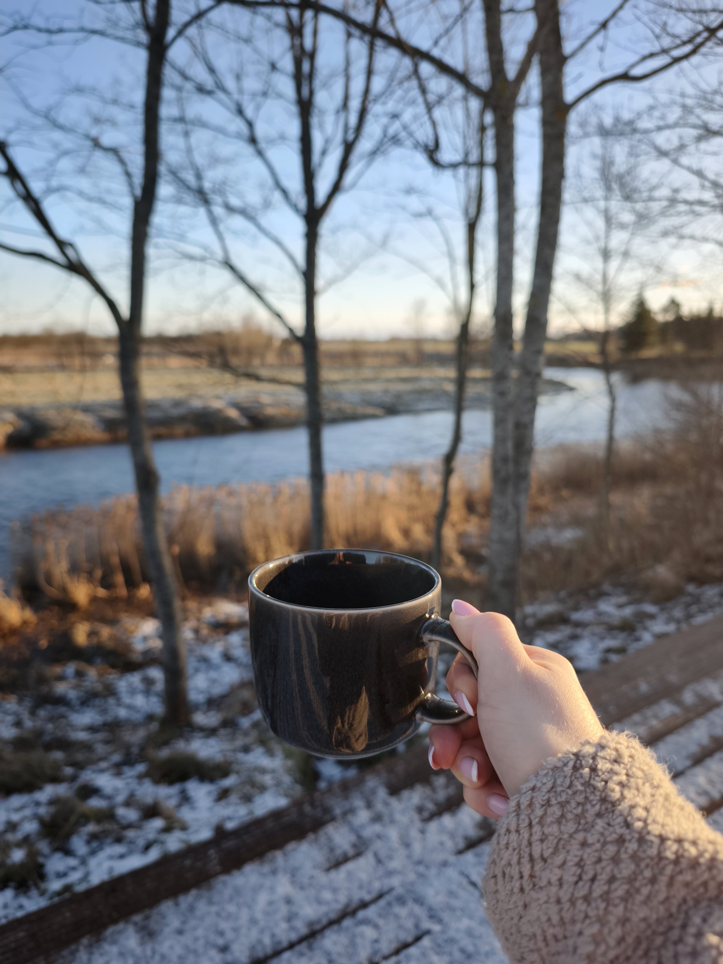 Person holding a black coffee mug outdoors near a river with leafless trees and snow on the ground during sunset or sunrise.