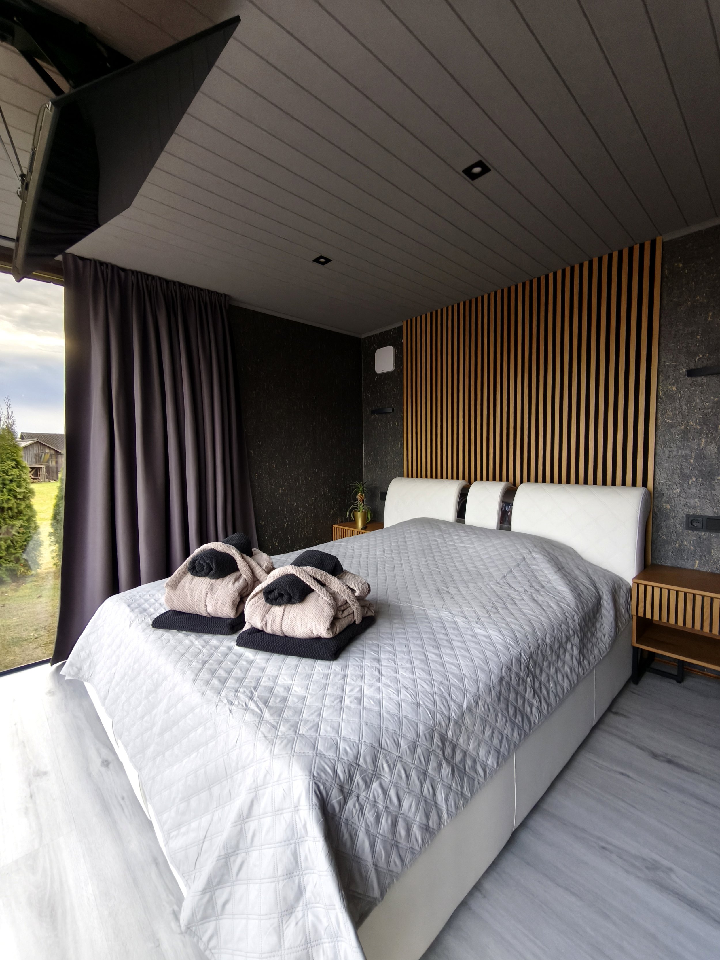 Modern bedroom with a white bed, folded towels with black and beige colors on the bed, gray quilted bedspread, dark curtain, wood paneling on the wall behind the bed, and a large window showing an outdoor landscape.