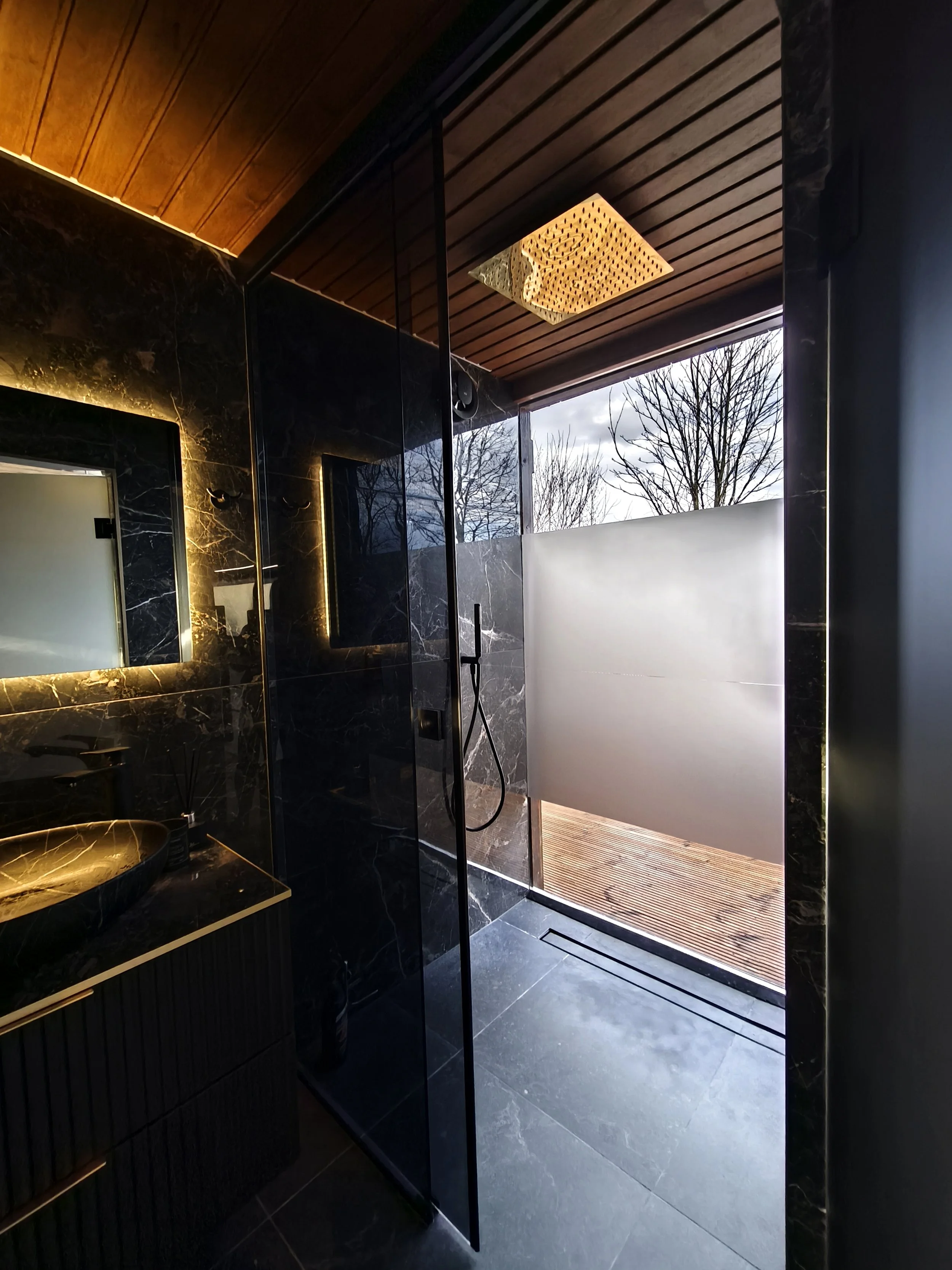 Modern bathroom with black marble walls, a black marble sink, a mirror with backlit frame, a wood-paneled ceiling, a frosted glass door, and a large window showing outdoor trees.
