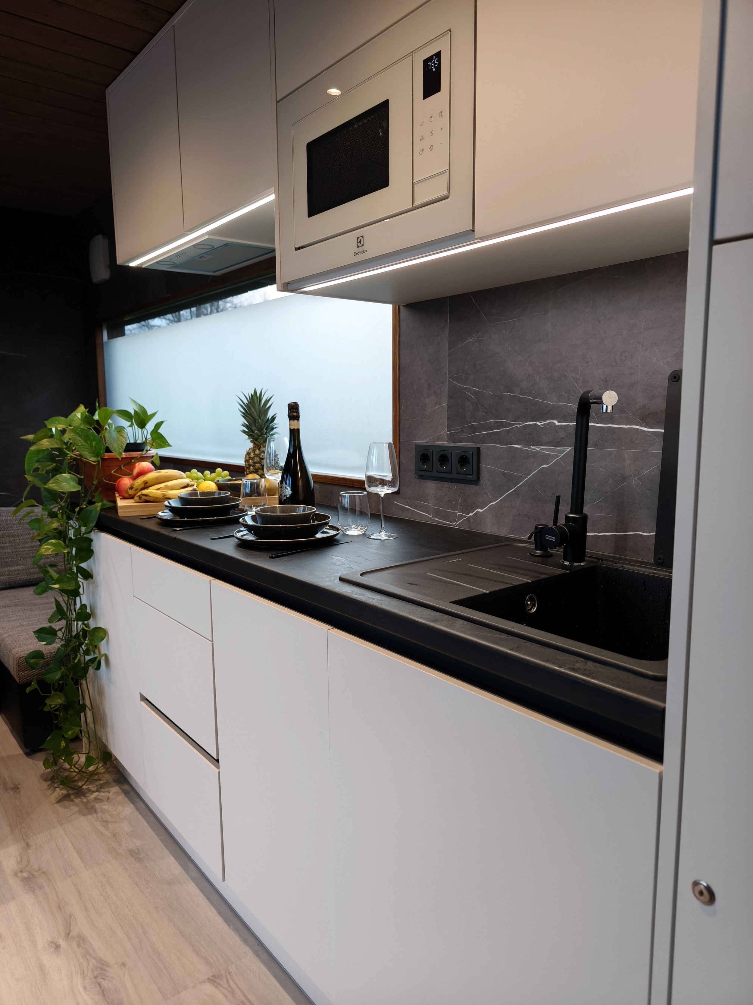 Modern kitchen with white cabinets, black countertop, dark marble backsplash, and black sink with faucet. There are fruits, wine, glasses, and a plant on the counter, with a frosted window in the background.
