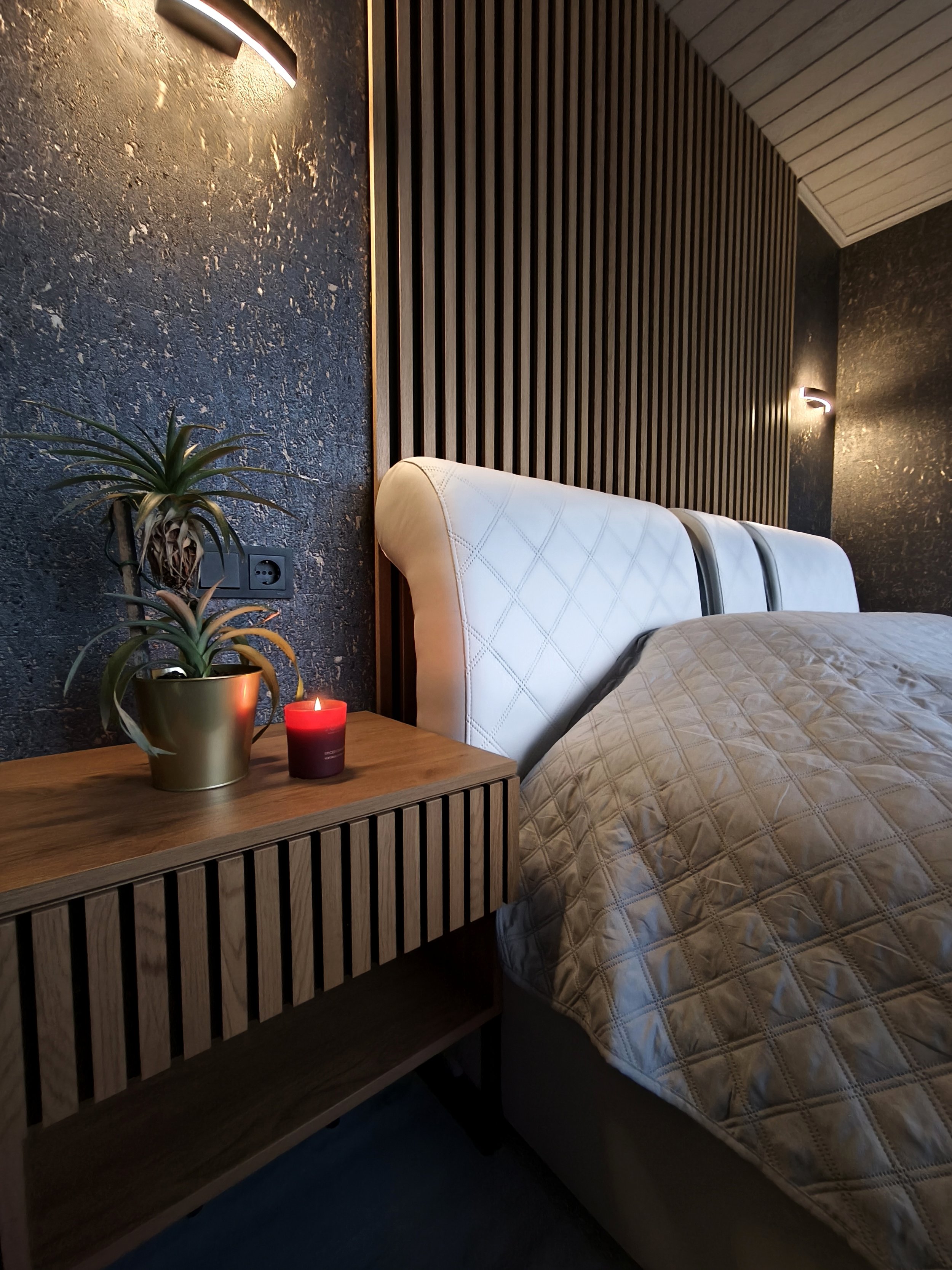 A bedroom corner with a wooden nightstand, a potted plant, a lit red candle, a textured beige bedspread, a white upholstered bed headboard, dark textured accent wall, and wall-mounted lights