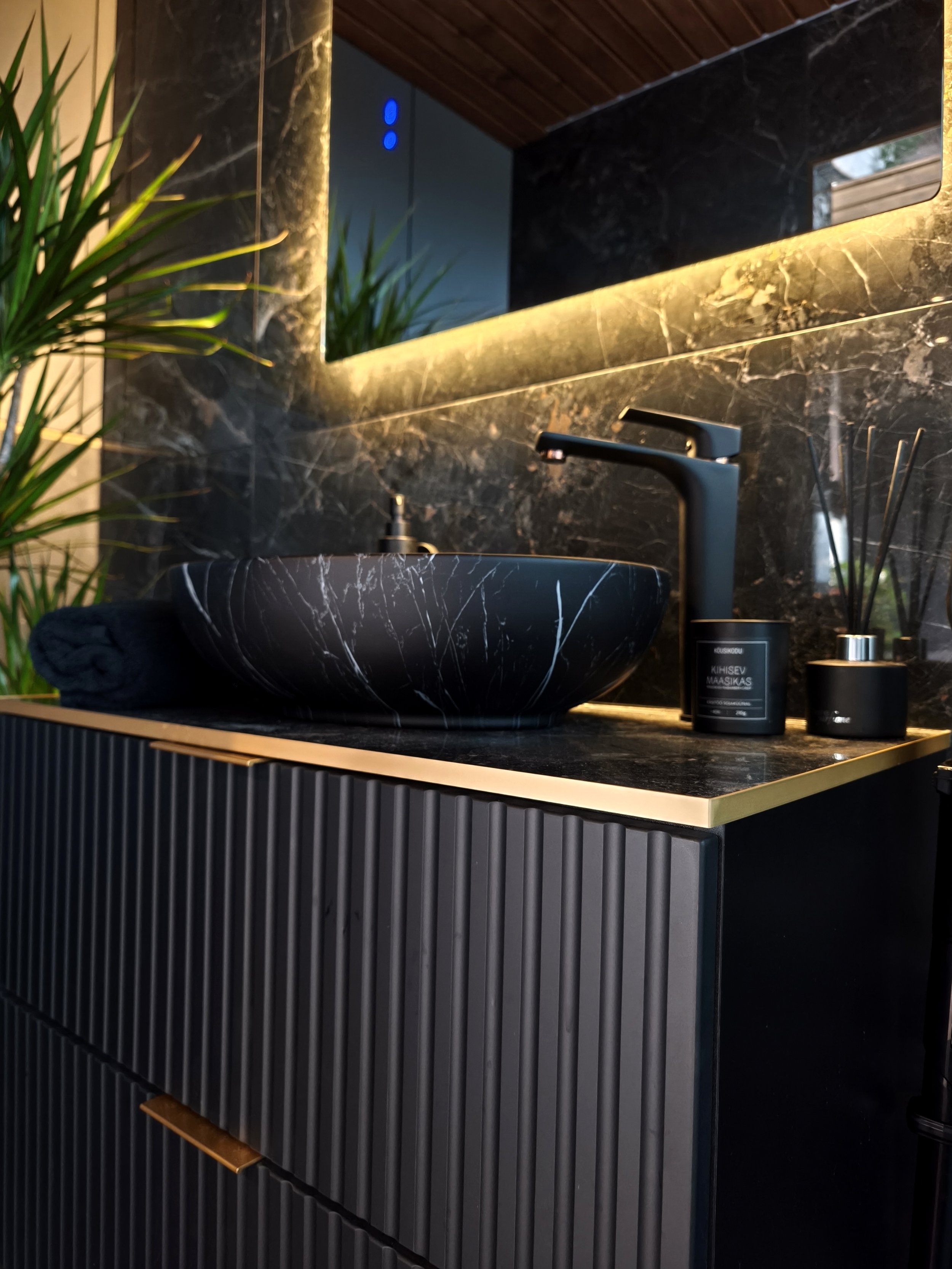 Modern bathroom sink with black marble vessel basin, black faucet, and black accessories on a black and gold vanity next to a mirror with backlighting, with plants reflected in the mirror.