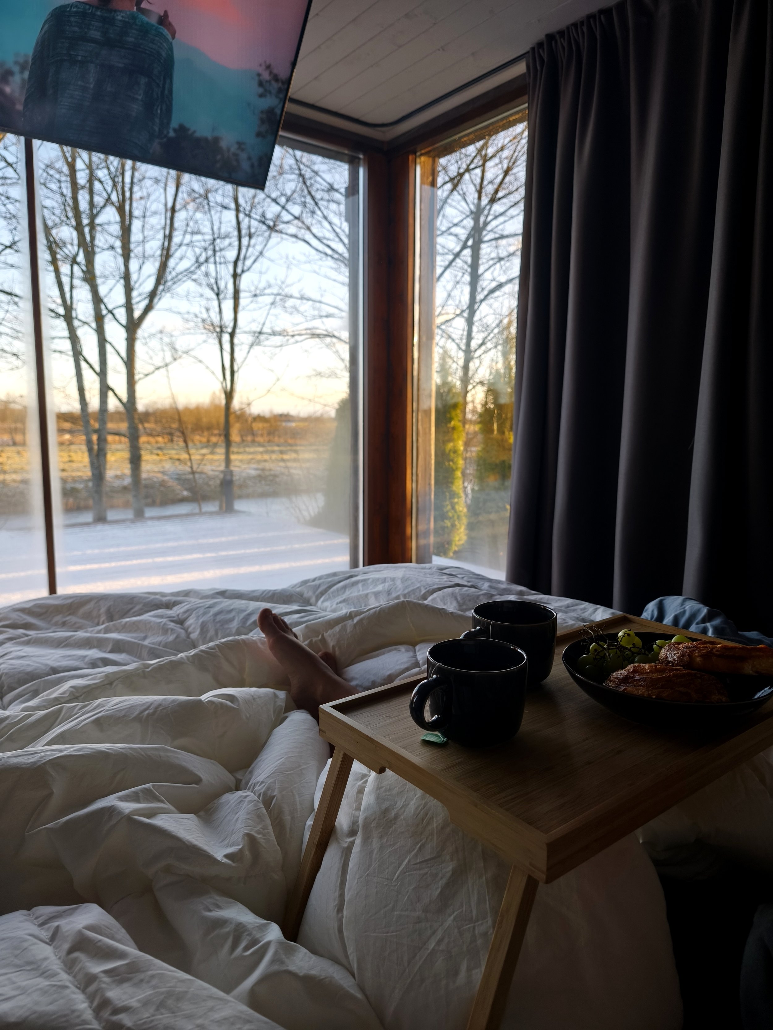 A cozy bedroom with large windows showing a winter landscape outside. A person is lying in bed with white bedding, and a wooden tray with two black coffee mugs, a bowl of grapes, and some food is resting on their legs.