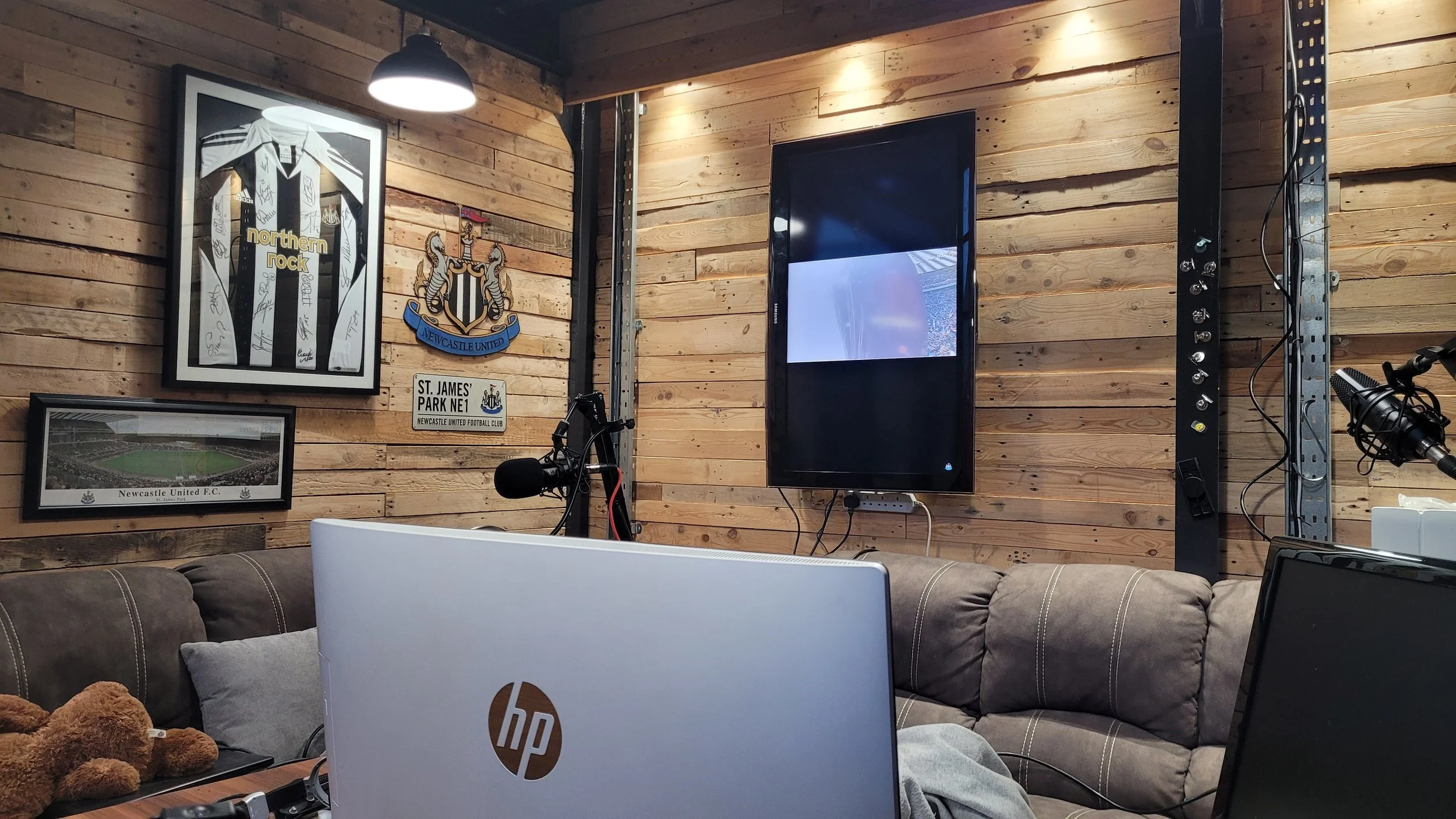 A cozy room with wooden walls decorated with sports memorabilia, including Newcastle United FC posters and a Newcastle United crest. There is a big television on the wall, a microphone, and various electronic equipment. A couch with pillows and a computer with an HP logo are visible in the foreground.