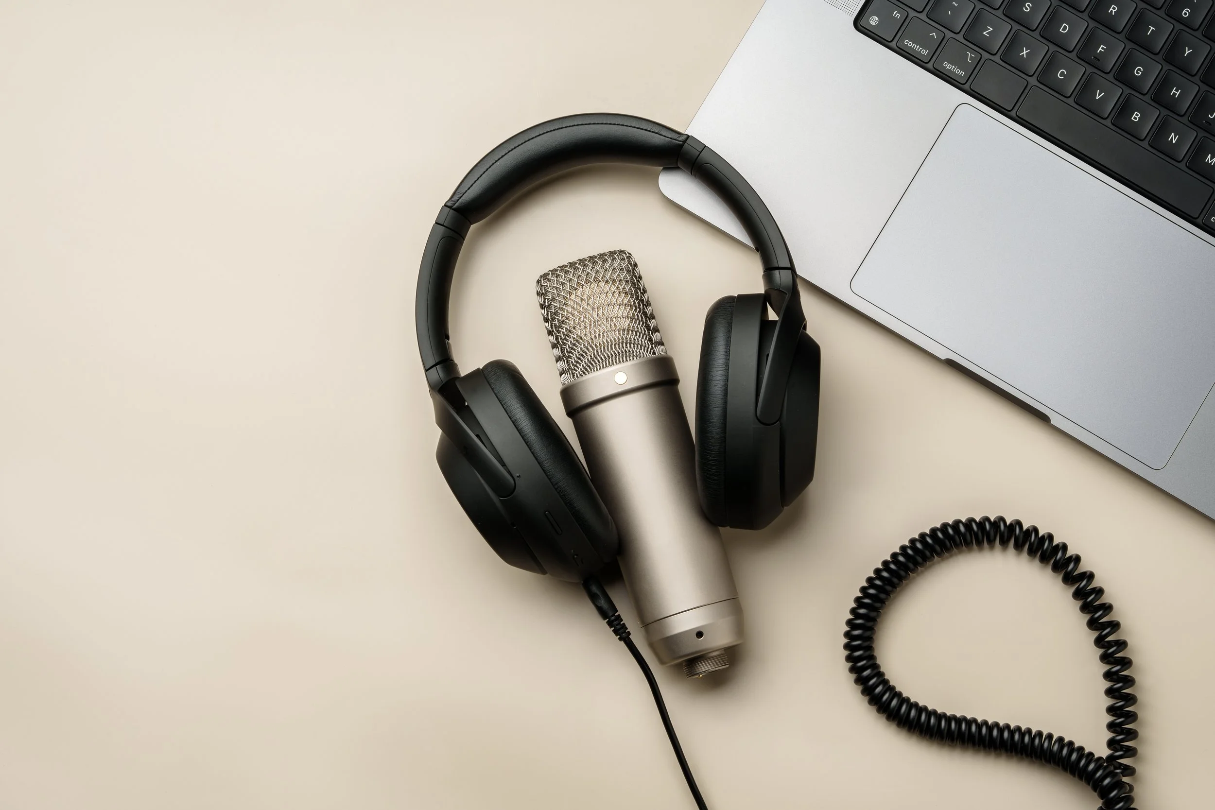 A pair of black over-ear headphones, a silver microphone, a silver laptop, and a coiled black cable on a light-colored desk.