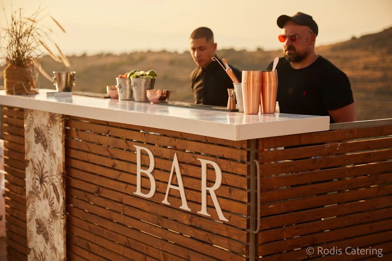 An outdoor bar setup with two men behind the counter during sunset, one wearing a black shirt and sunglasses, the other in a black T-shirt. The bar has wood slats with a sign reading 'BAR' and various bar tools and decorations on top.