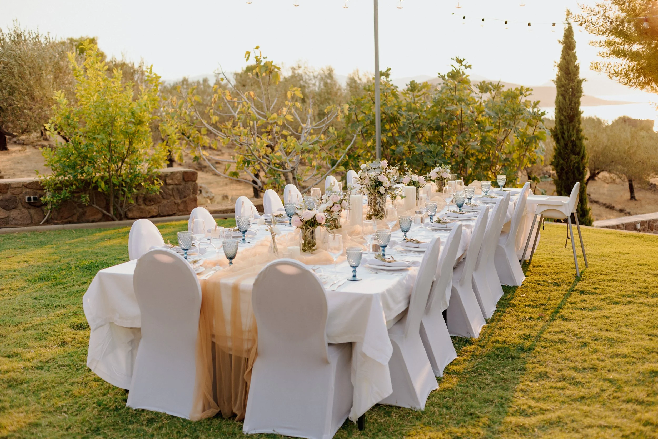 Elegant outdoor banquet table set with white tablecloths, floral centerpieces, glassware, and place settings on a lush green lawn, with a scenic backdrop of trees and a sunset sky.