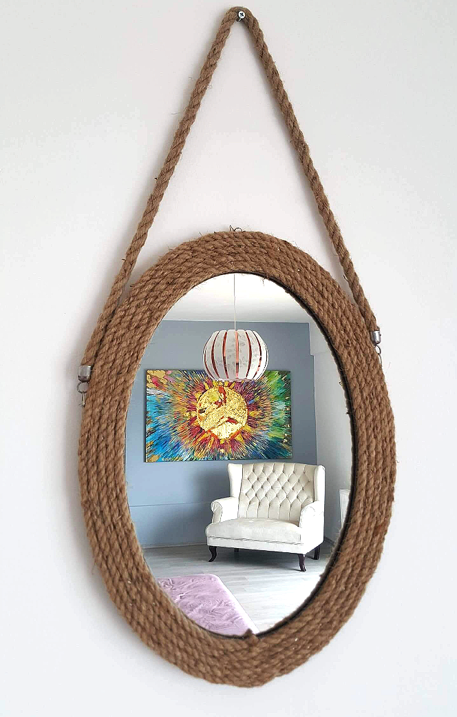 A mirror with a brown rope frame hanging on a wall, reflecting a living room with a white tufted armchair, a colorful abstract painting, a pink rug, and a spherical hanging lamp.