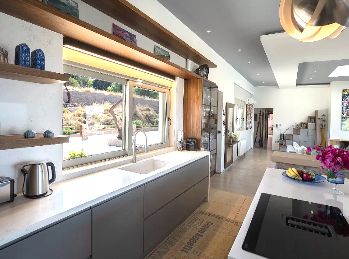 Bright kitchen with large window overlooking outdoor landscape, featuring modern gray cabinetry, white countertop, stainless steel appliances, wooden shelves with decorative items, and a dining area with a fruit centerpiece and flowers.