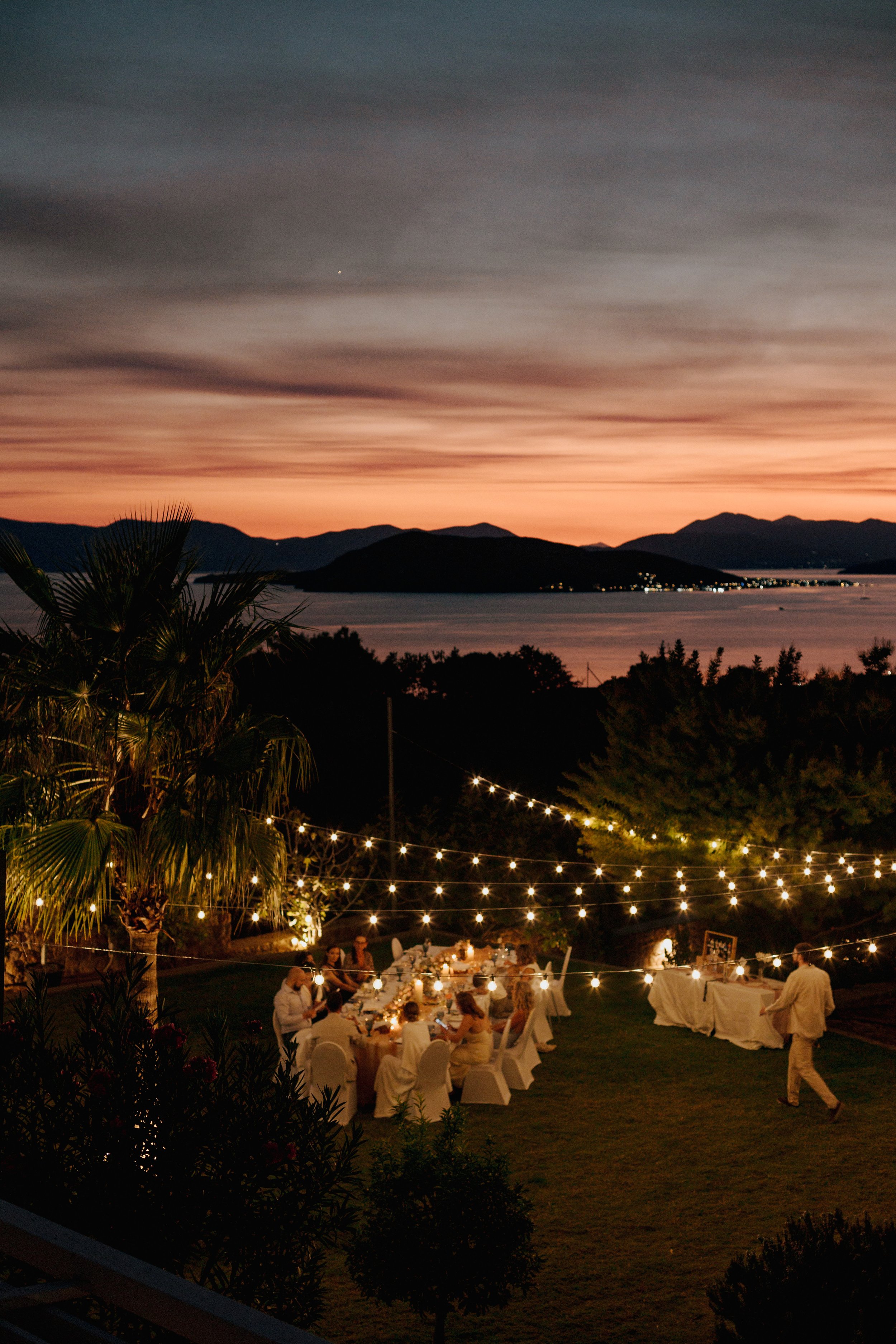 Outdoor evening event with a long dinner table, string lights, and guests, overlooking a body of water and mountain range at sunset.