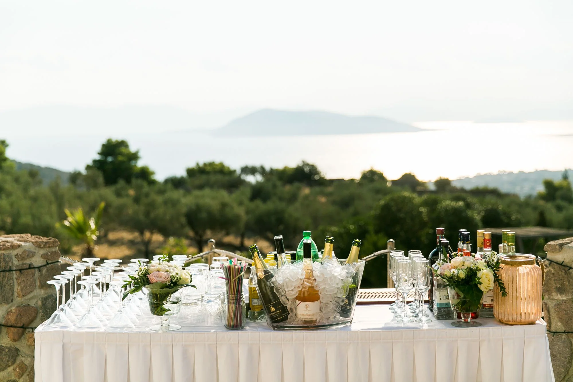 Outdoor bar setup with various bottles, glasses, straws, and floral arrangements on a white tablecloth, overlooking a scenic landscape with trees and water in the background.