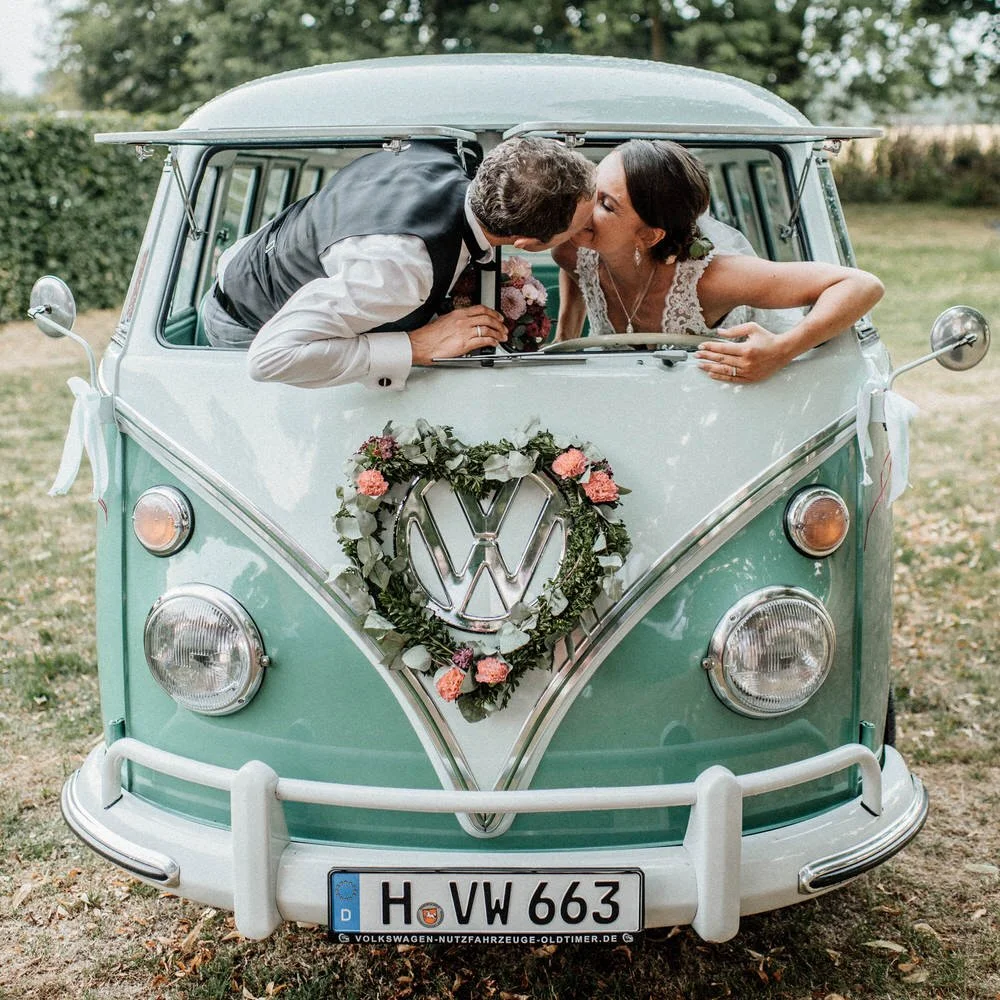 A bride and groom kissing through the open front window of a vintage Volkswagen van decorated with a floral wreath and a Volkswagen logo for their wedding celebration.