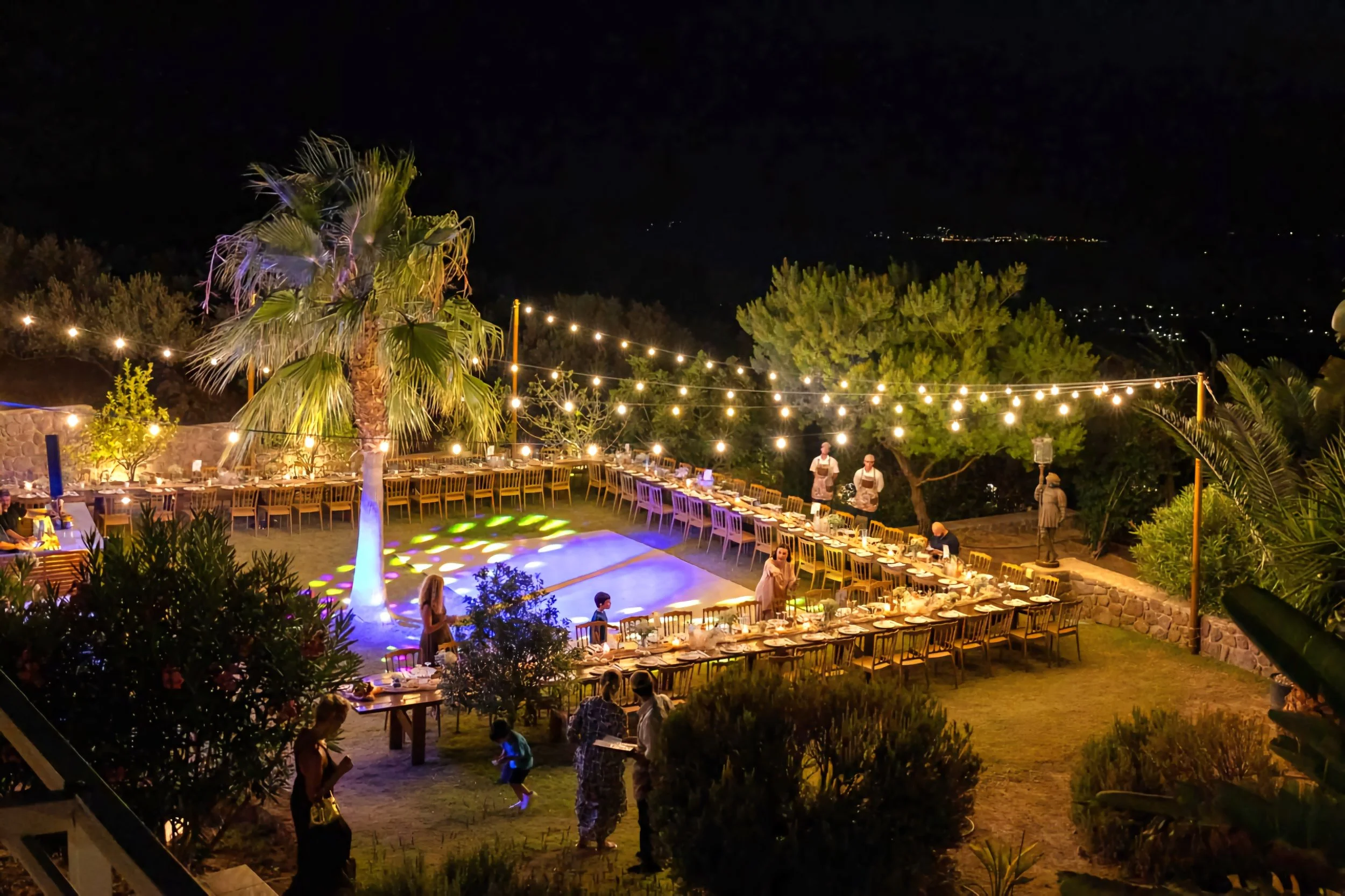 Night outdoor event with a long dining table set under string lights, surrounding a lit palm tree and trees, with people, some children, engaging in activities.