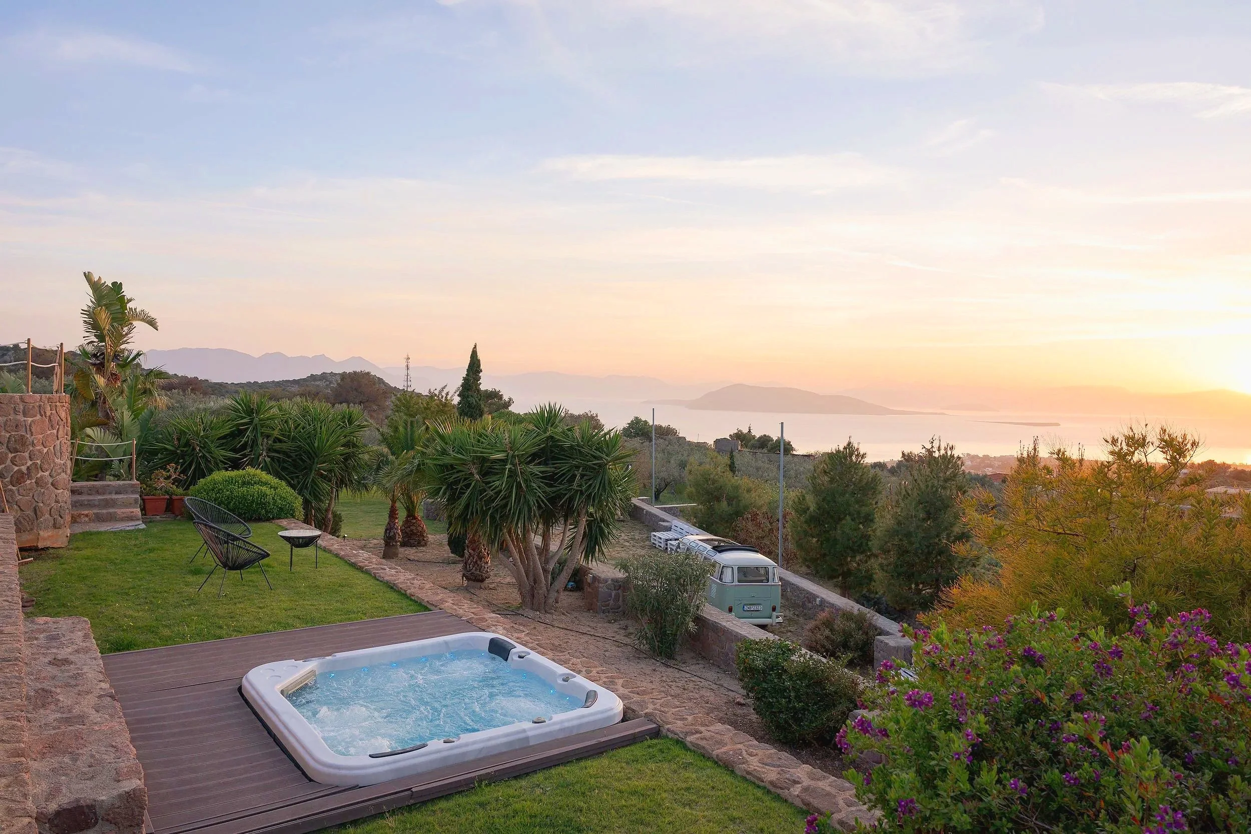 A backyard with a hot tub on a deck, surrounded by green grass, plants, and outdoor chairs, overlooking a scenic landscape with trees, hills, and water during sunset.