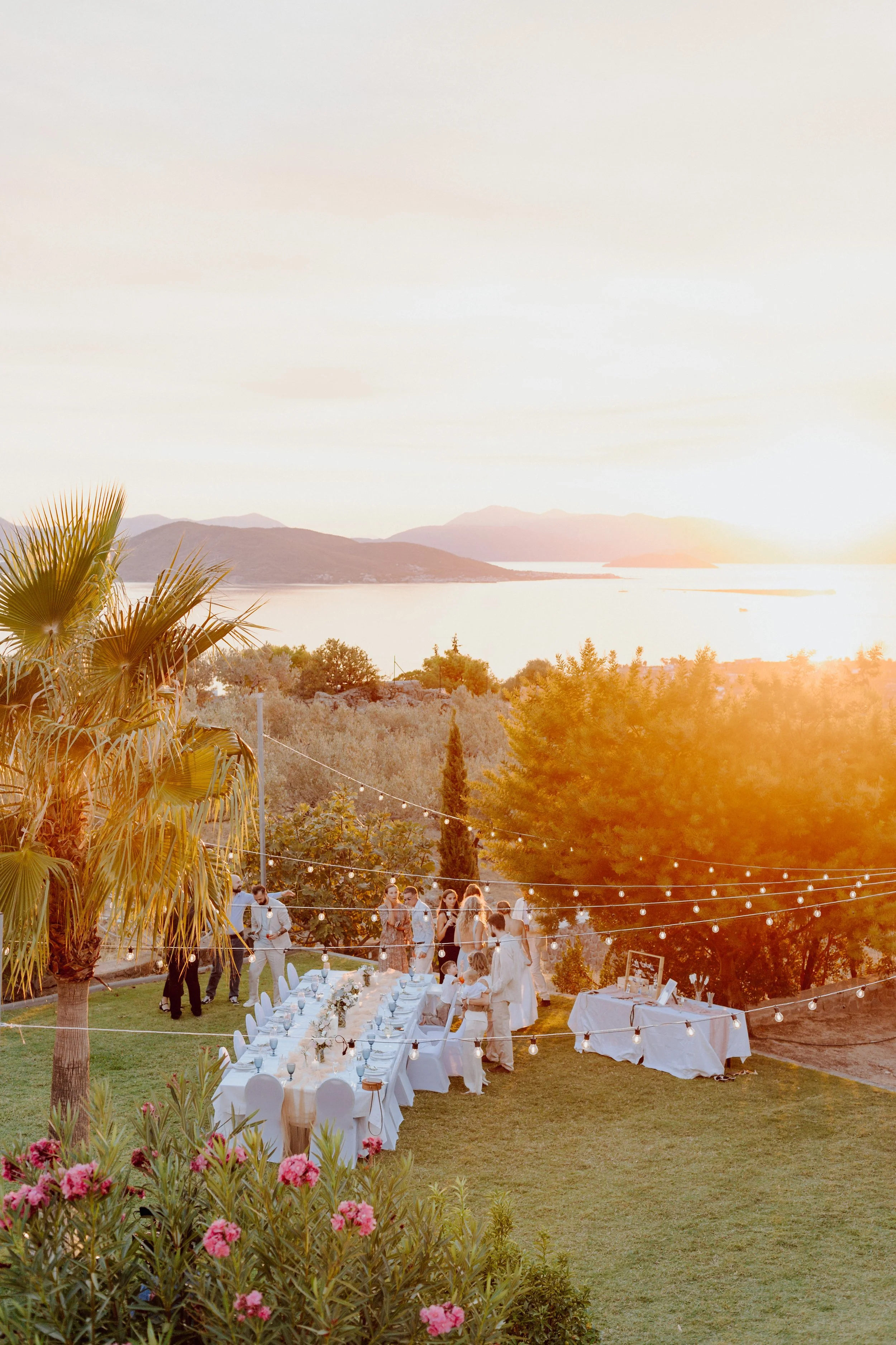 Outdoor dinner party at sunset with a long table, string lights, guests, and scenic view of water and mountains in the background.