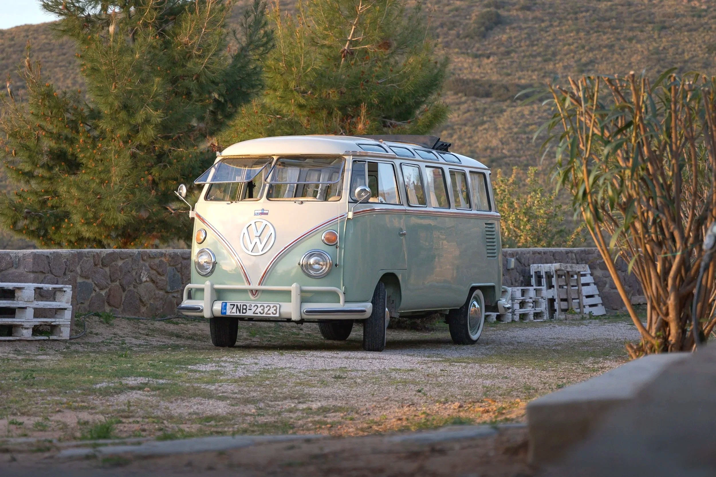A vintage light green Volkswagen Type 2 van parked outdoors near trees and a stone wall.