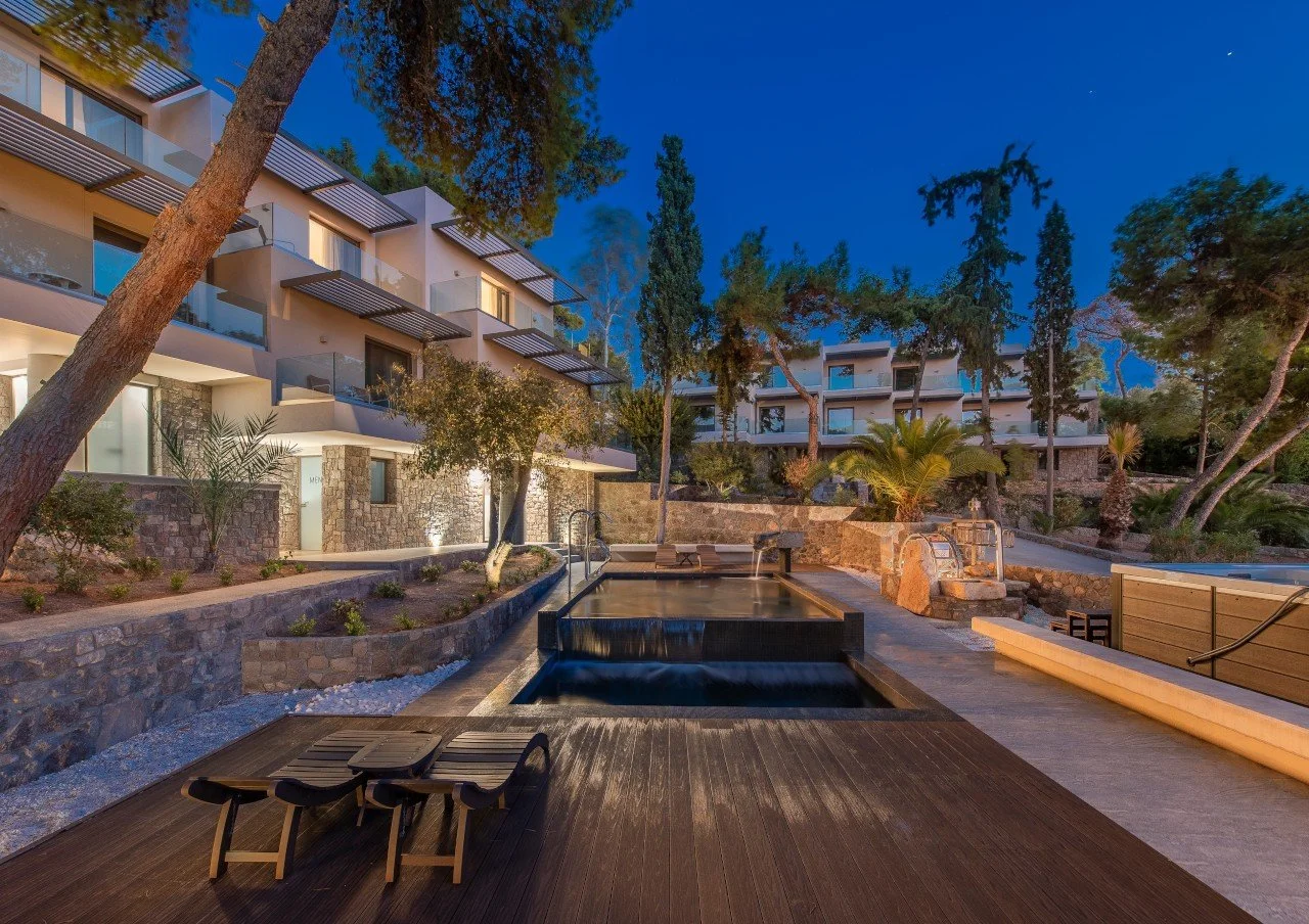 Luxury apartment complex with outdoor pool and hot tub, surrounded by trees and landscaped gardens, in twilight.